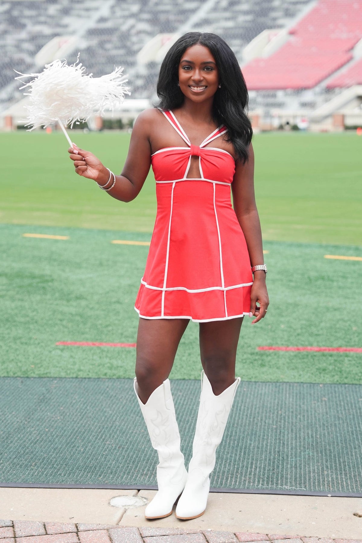 A woman stands on a football field wearing a bright red and white cheerleader-style mini dress with white cowboy boots, holding a white feather fan.