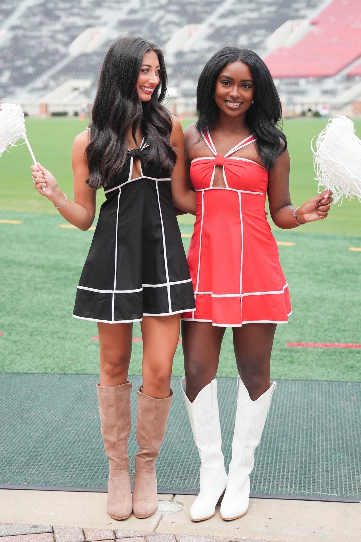 Two women posing on a football field in cheerleader-style mini dresses. One dress is black with white piping and tan knee-high boots, and the other is red with white piping and white cowboy boots.