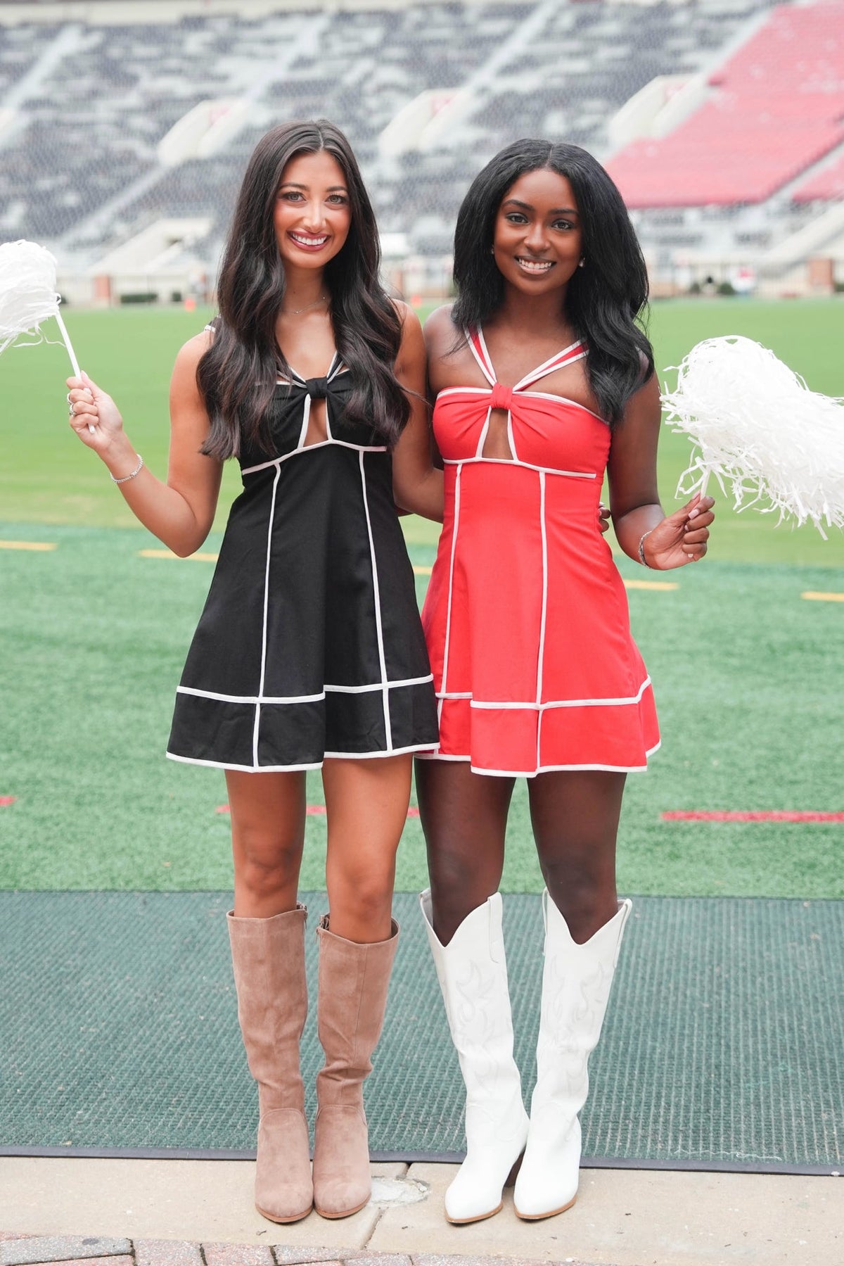 Two women posing in matching cheerleader-style mini dresses, one black with white trim and one red with white trim, standing on a football field with stadium seating in the background.