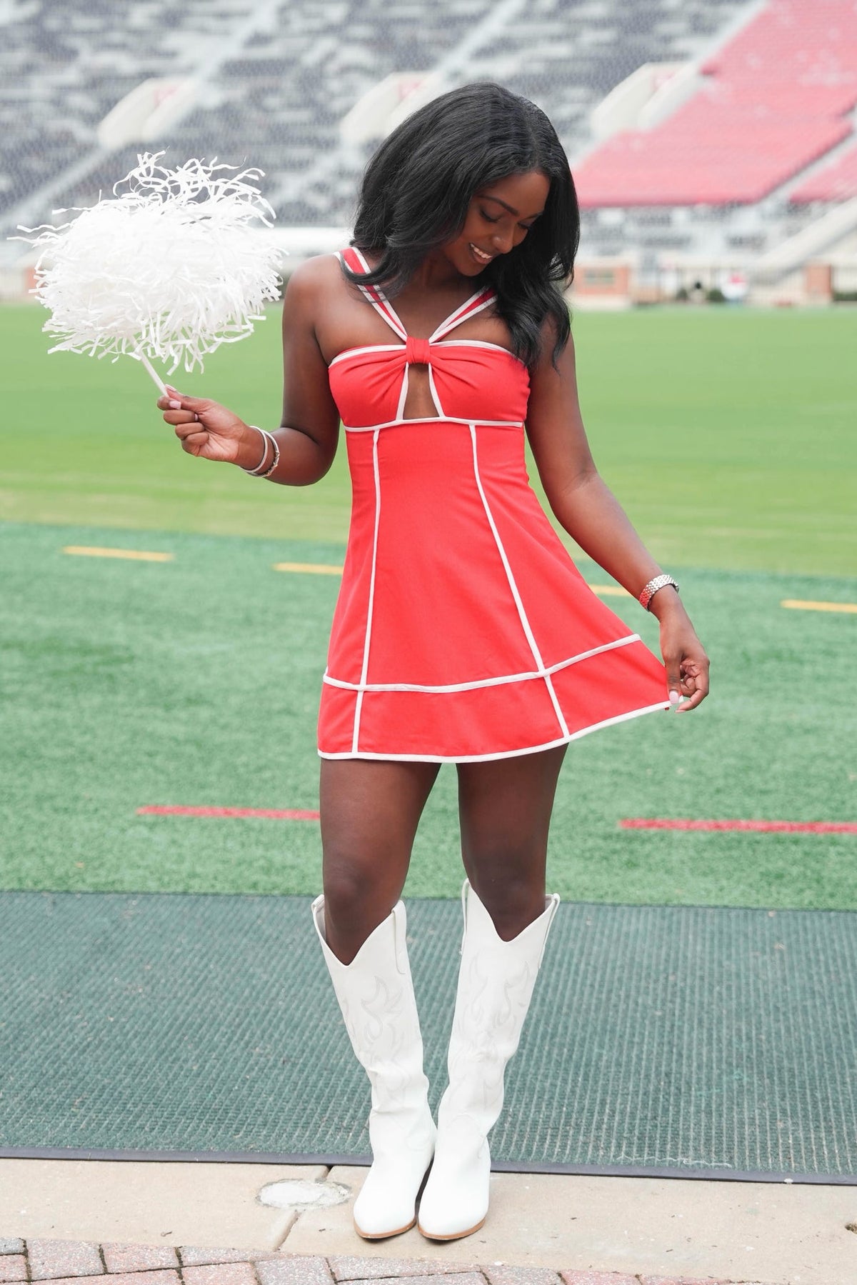 A woman stands on a sports field wearing a bright red mini dress with white trim and white cowboy boots, holding a white feather pom-pom. The background shows stadium seating and green turf.