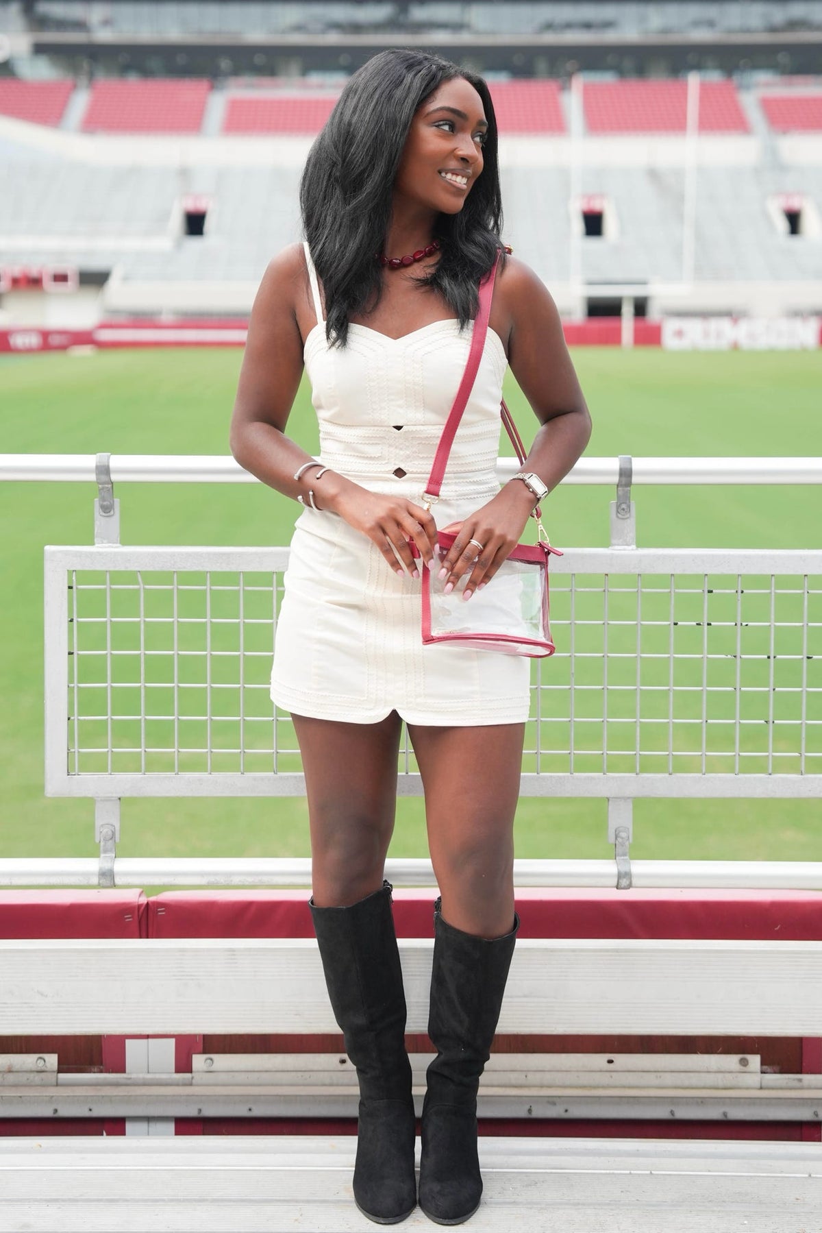 A woman stands in a stadium setting wearing a white sleeveless mini dress with black knee-high boots and holding a white crossbody bag with red trim.
