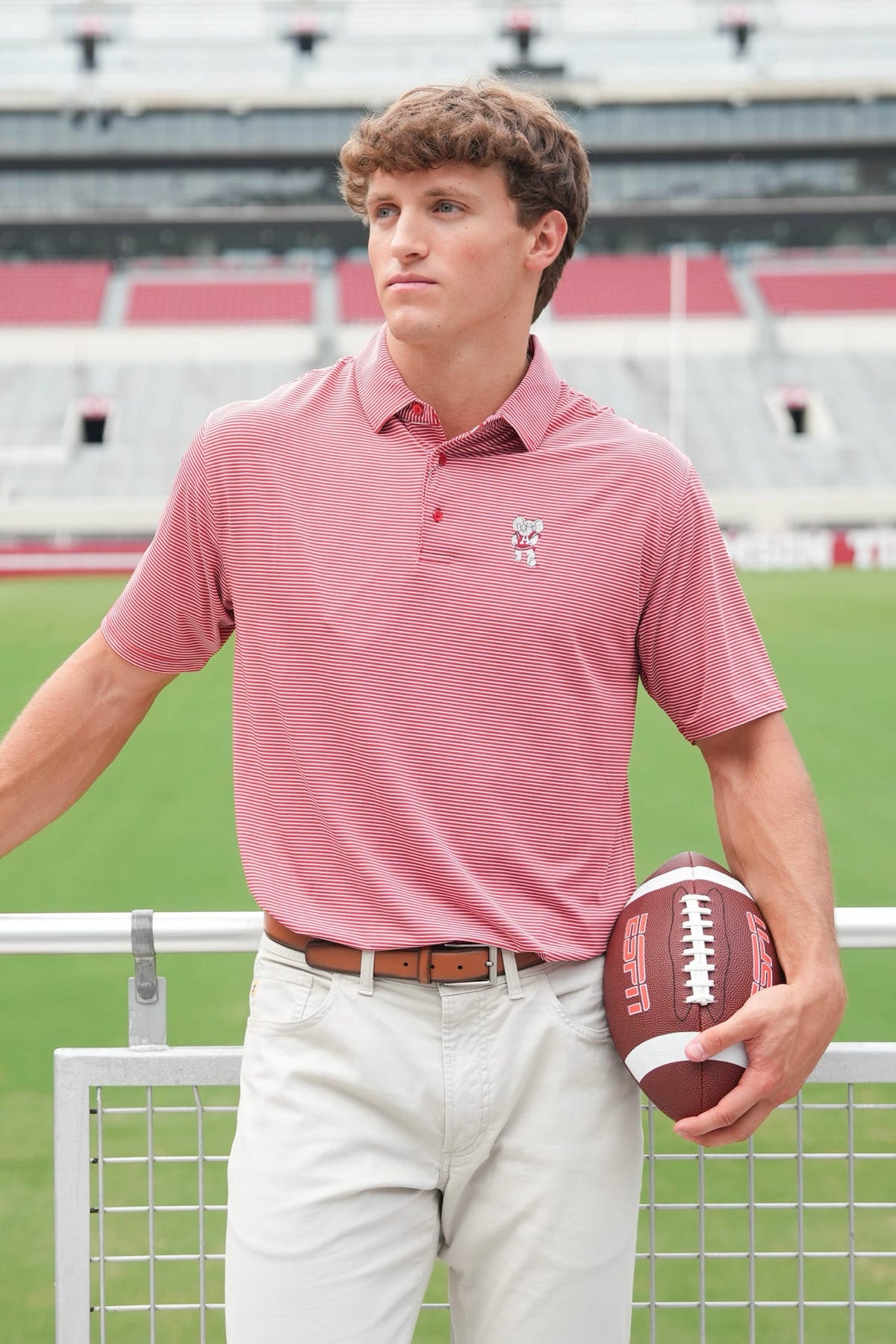 A man wearing a pink polo shirt and khaki pants stands on a football field holding a football, with stadium seating in the background.