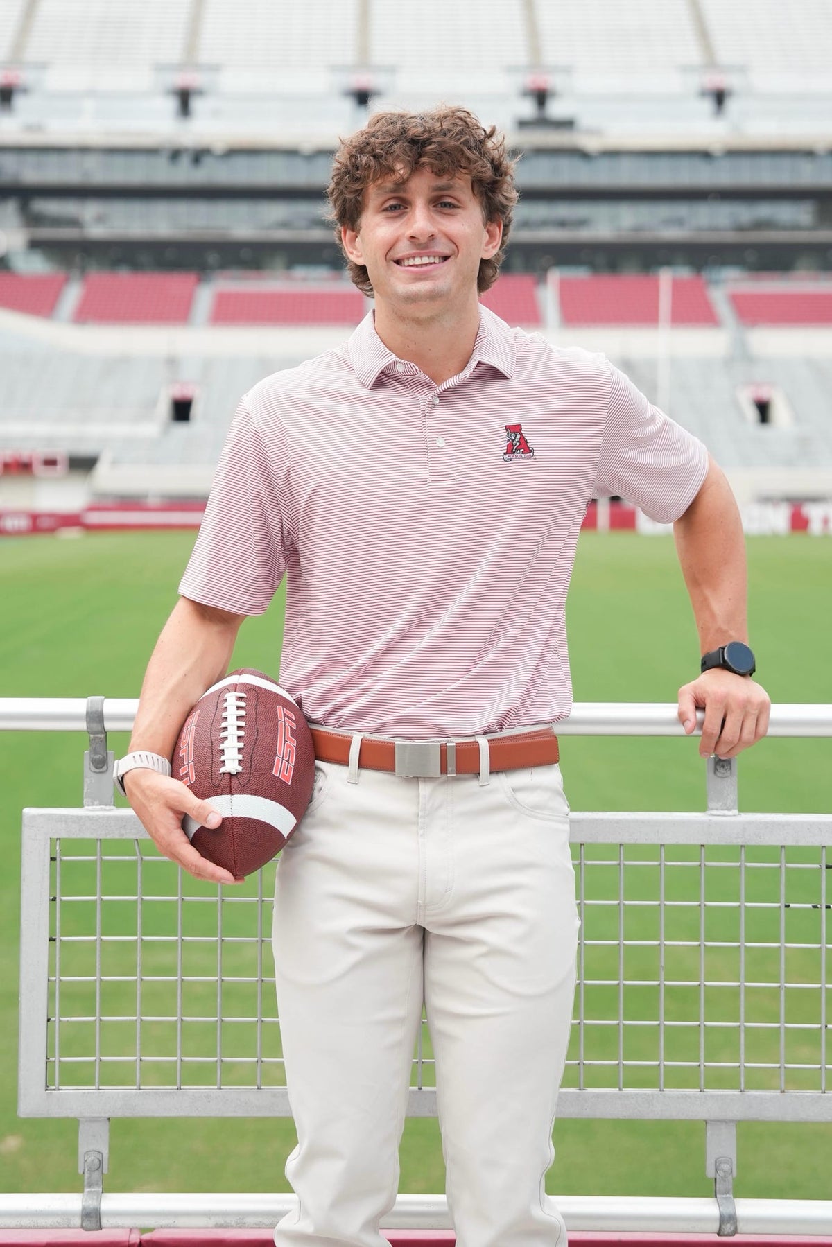 A man stands in a stadium holding a football, wearing a light pink polo shirt with a small red logo on the chest, white pants, and a brown belt.
