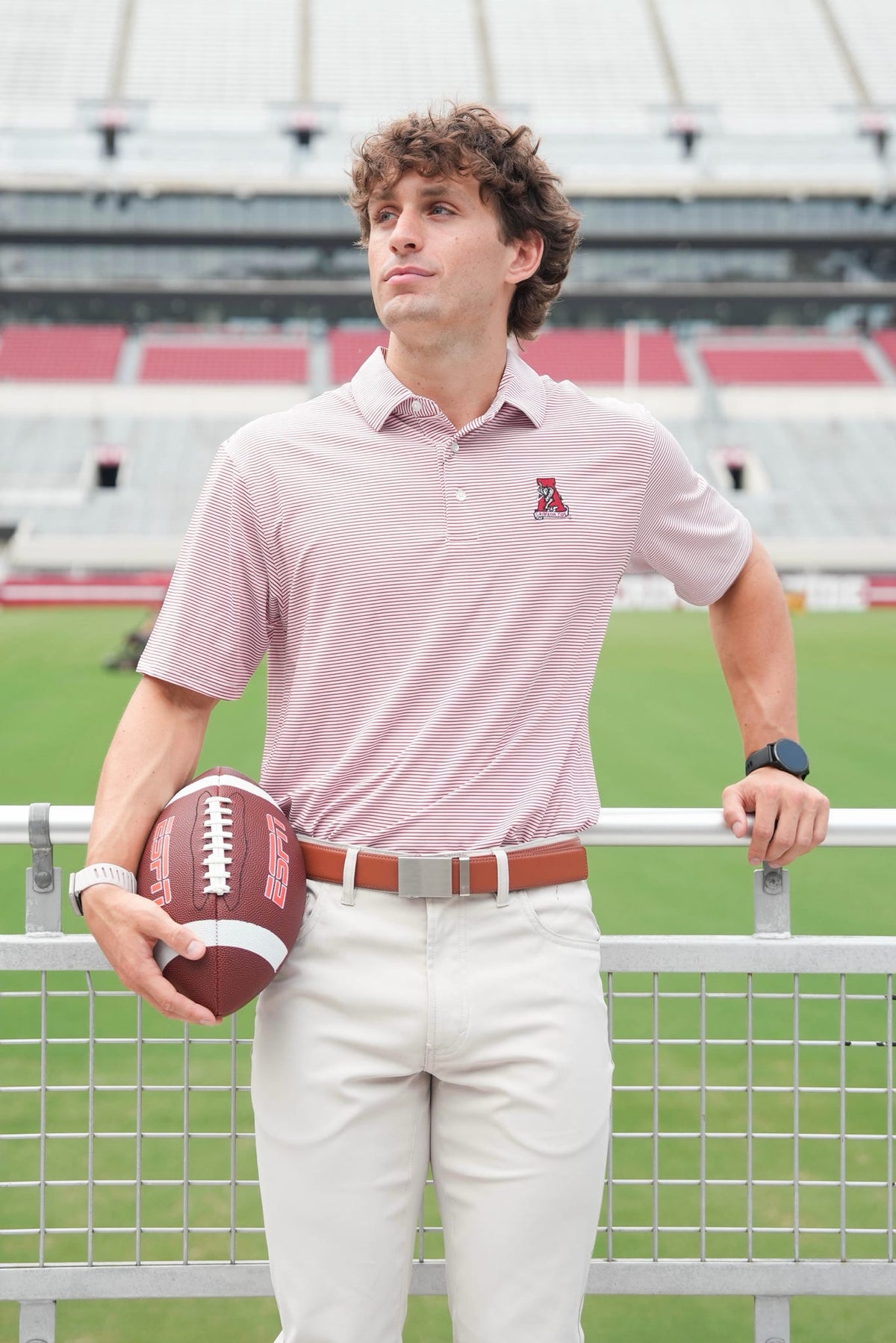 A man stands on a football field wearing a light pink polo shirt with a small red logo on the left chest, white pants, and holding a brown football.