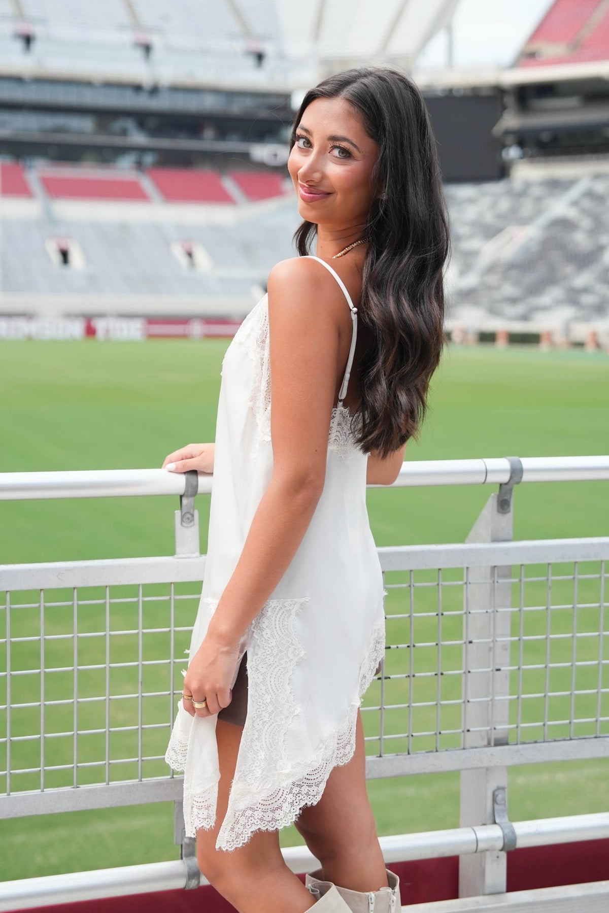 A woman with long dark hair stands on a stadium balcony wearing a white sleeveless lace dress, with a green field and stadium seating in the background.