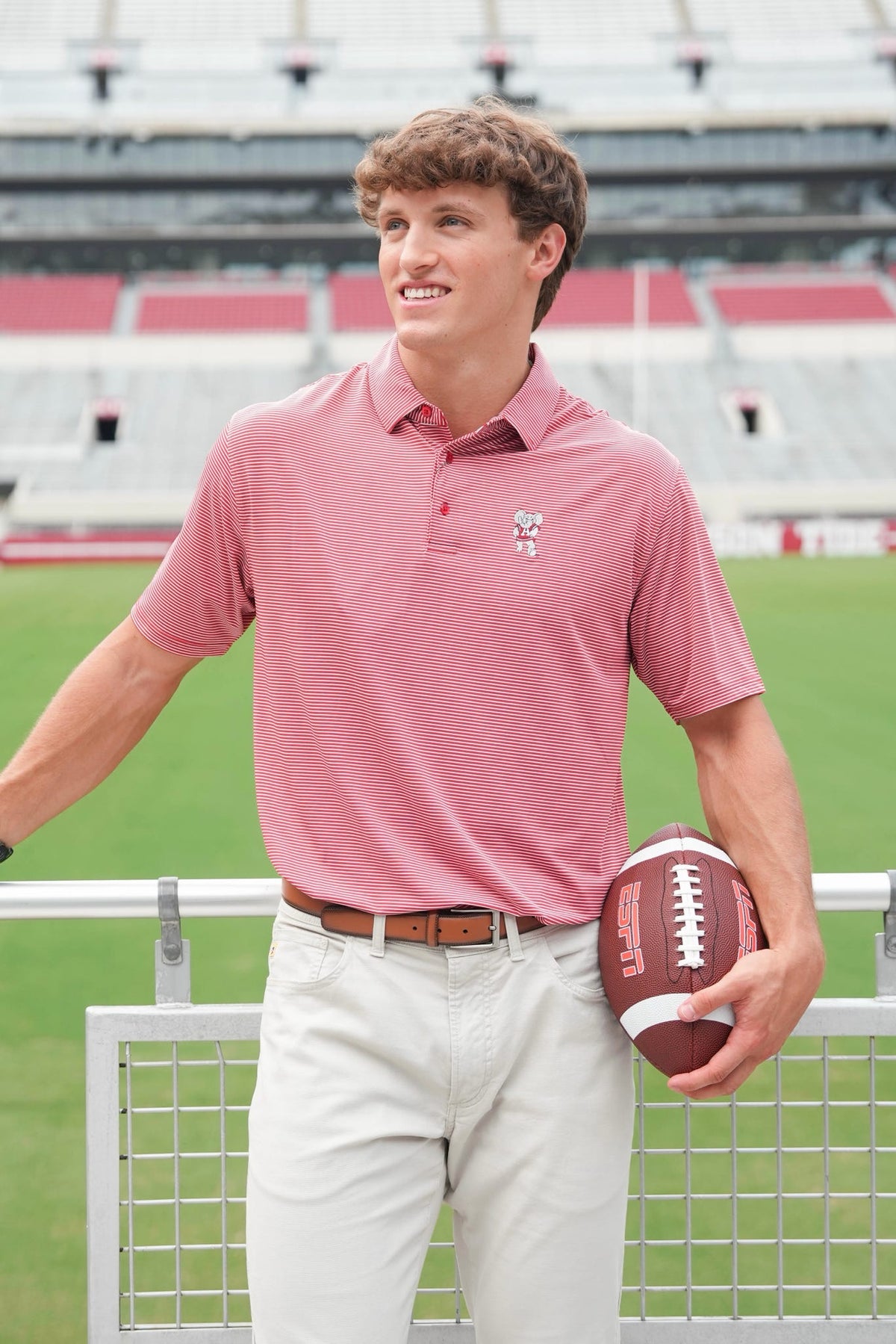 A man standing on a football field wearing a pink polo shirt with a small embroidered logo on the chest, paired with light khaki pants and holding a brown football.