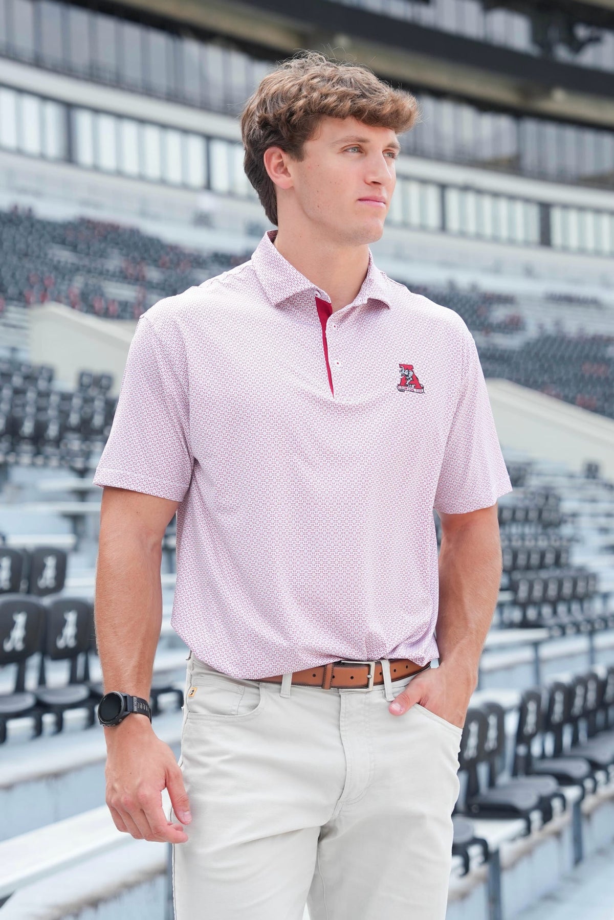 A man wearing a light pink micro-patterned polo shirt with a red inner collar and a small embroidered logo on the chest, paired with light beige pants, standing in front of stadium seating.