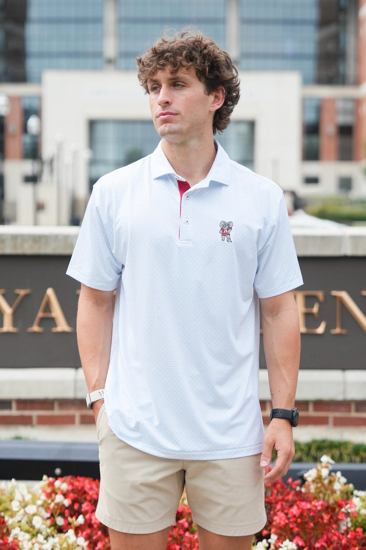 A man wearing a light blue polo shirt with a red inner collar and a small embroidered crest on the chest, paired with khaki shorts, standing outdoors in front of a modern building and flowers.