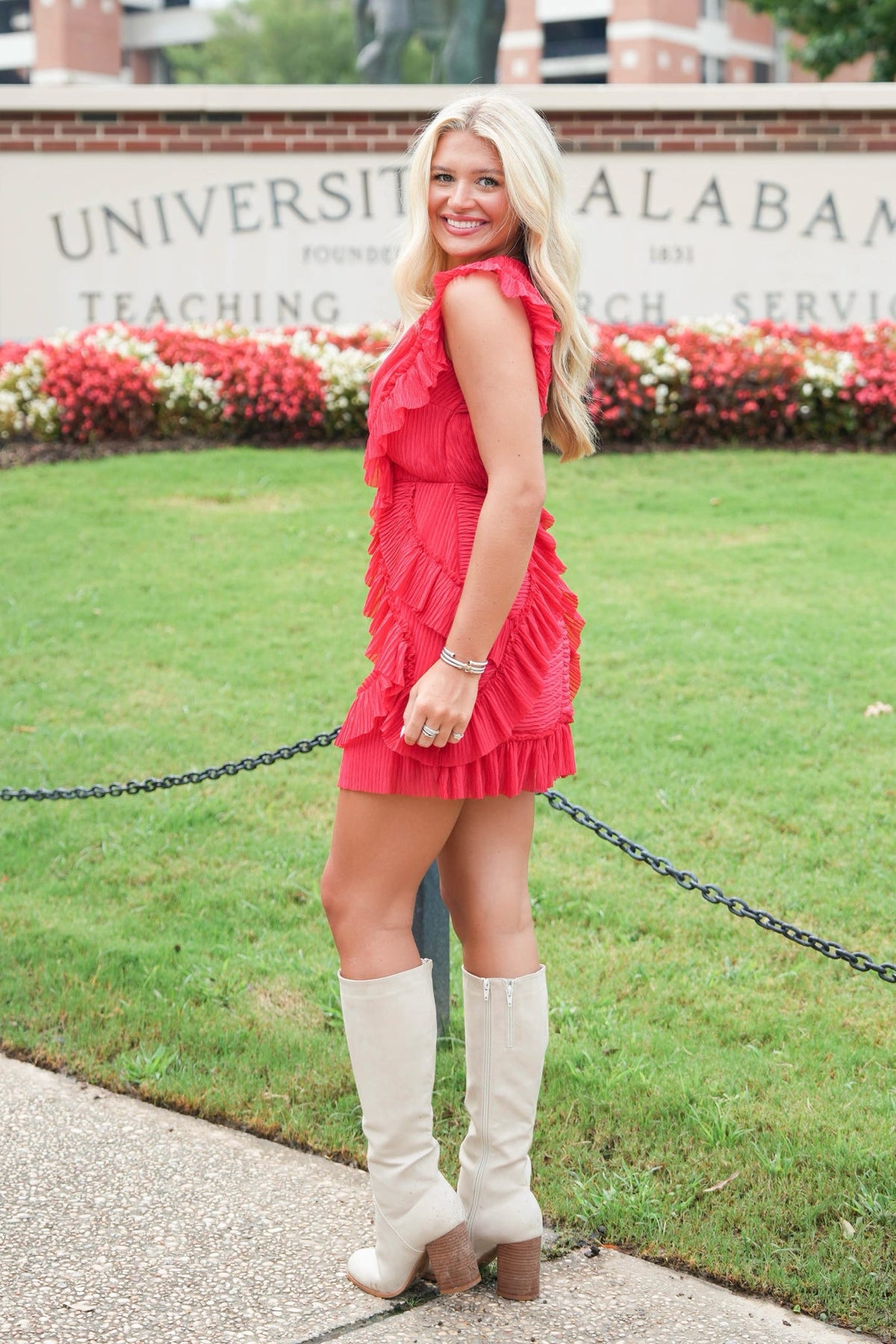 A woman posing outdoors in front of a University of Alabama sign, wearing a bright red ruffled mini dress and tall off-white knee-high boots.