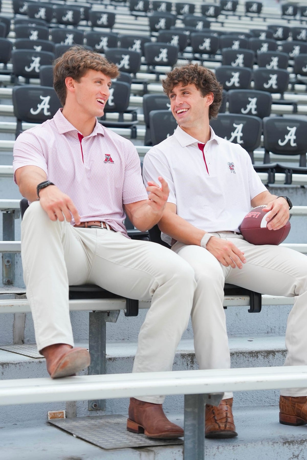 Two young men sitting on stadium bleachers wearing Alabama Crimson Tide athletic uniforms, with one holding a football. The background shows rows of stadium seats with the Alabama 'A' logo.