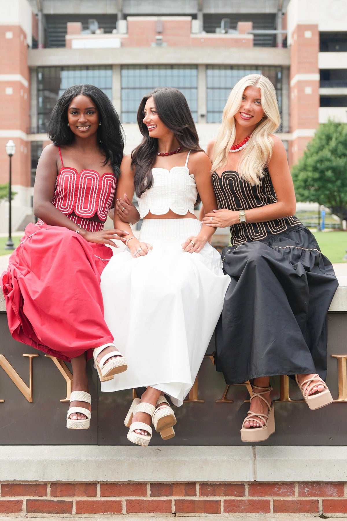 Three women sitting together outdoors on a brick ledge in front of a brick building. They are wearing coordinated outfits: one in a bright pink and red dress, one in a white two-piece set, and one in a black and gold dress.