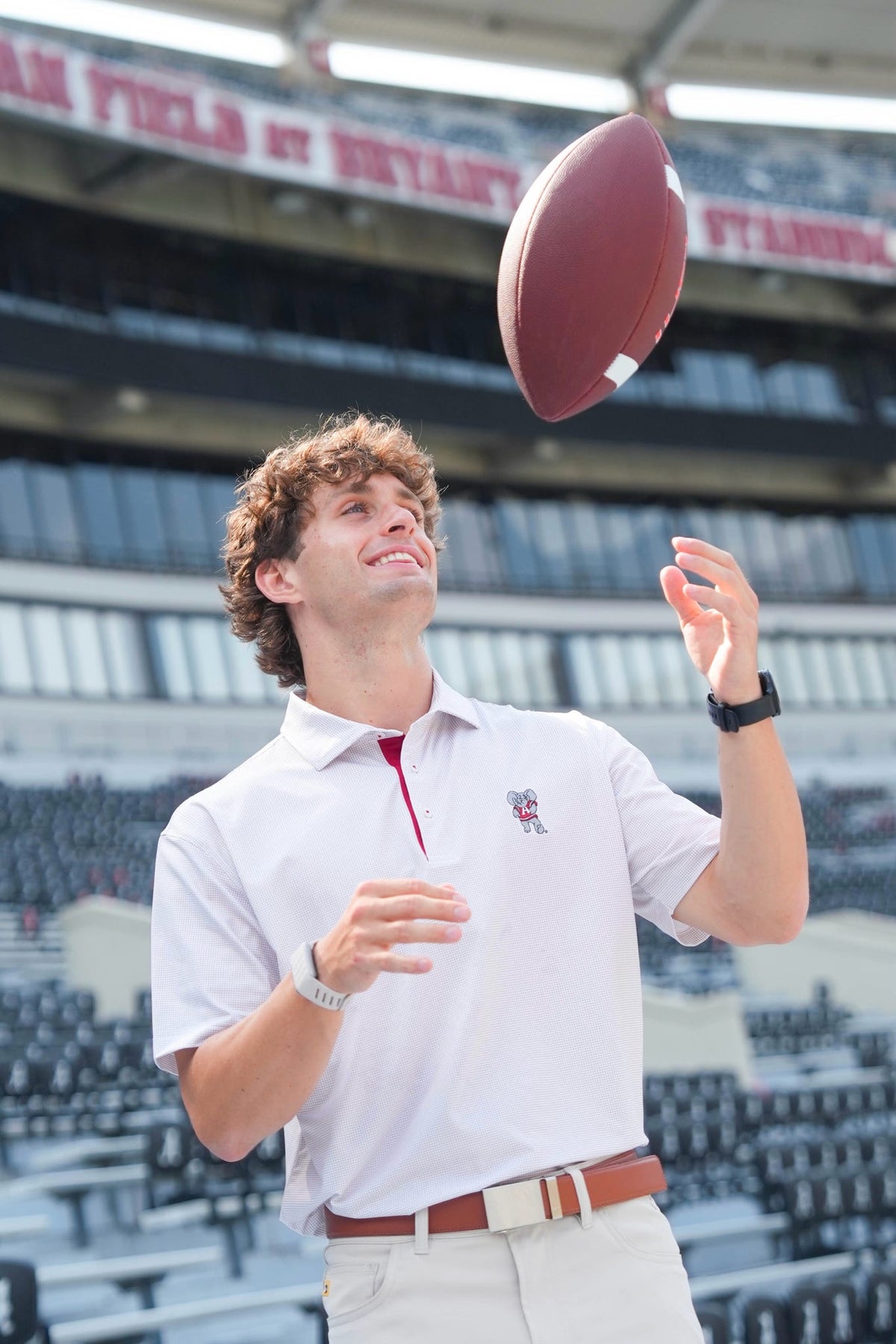 A man in a white polo shirt and khaki pants tosses a brown football in a stadium setting.