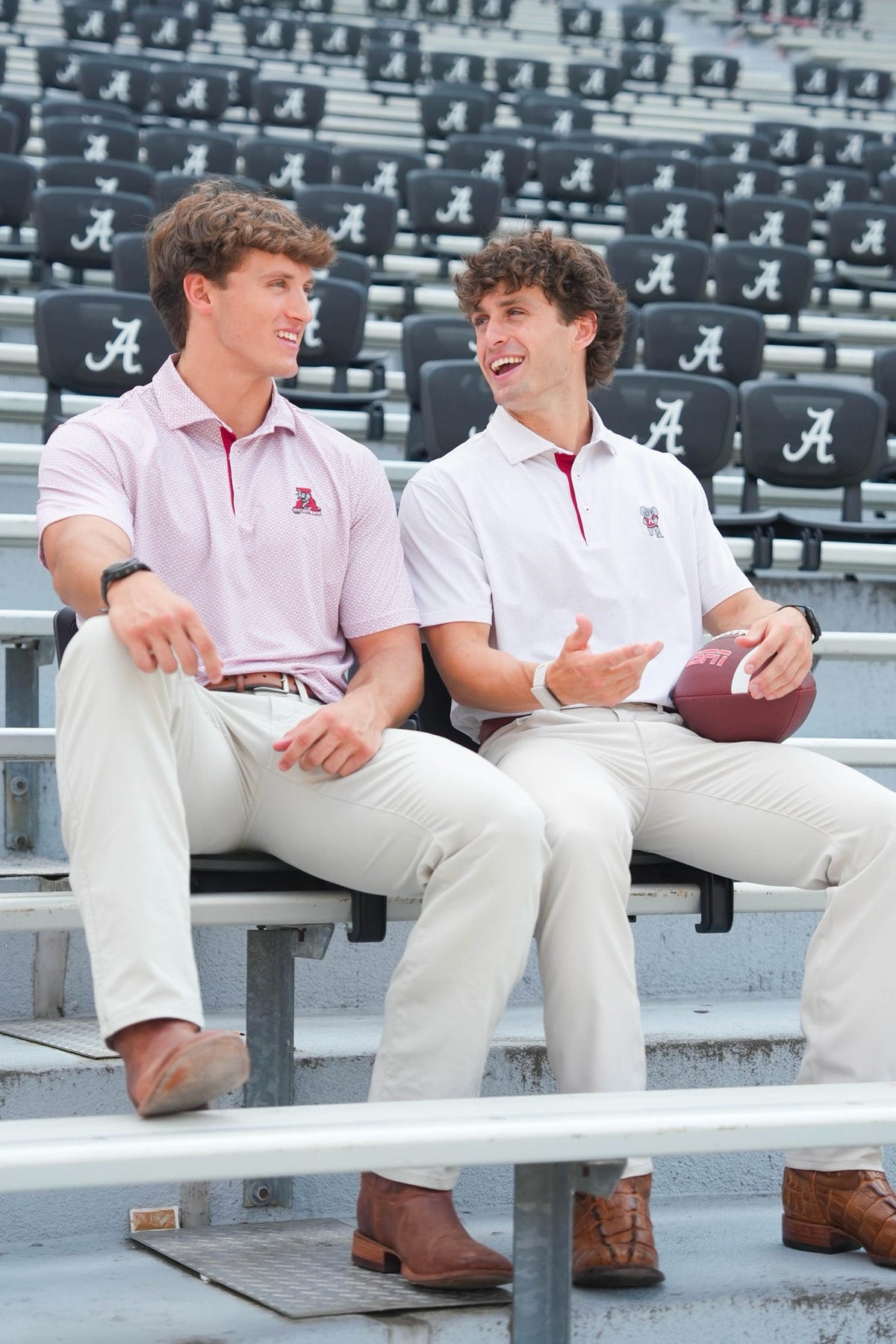 Two young men sitting on stadium bleachers wearing matching polo shirts and khaki pants, with Alabama-themed seats in the background.