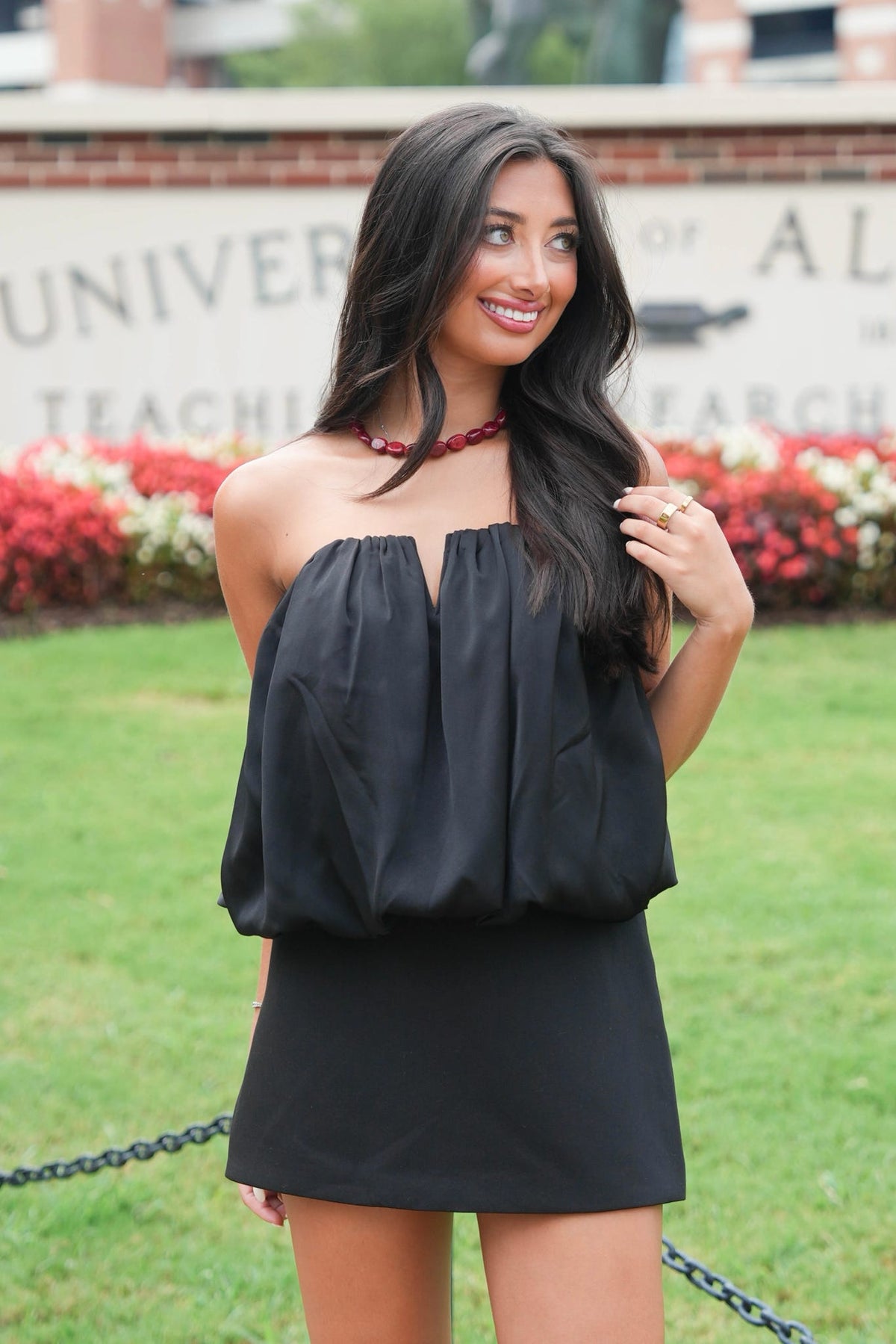 A woman modeling a black strapless mini dress with a gathered, balloon-style top and a straight skirt, standing outdoors in front of a grassy lawn and a brick sign.
