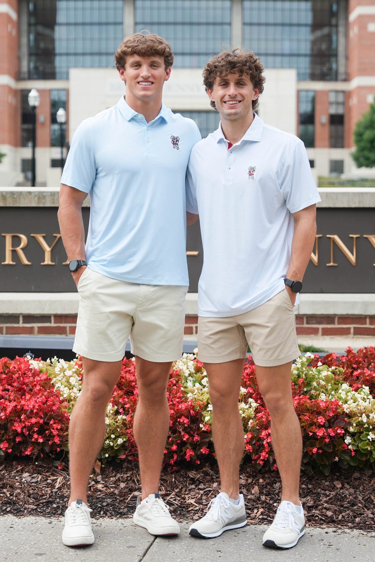 Two young men standing outdoors in front of a brick building and flowerbeds, wearing matching light blue polo shirts and beige shorts.