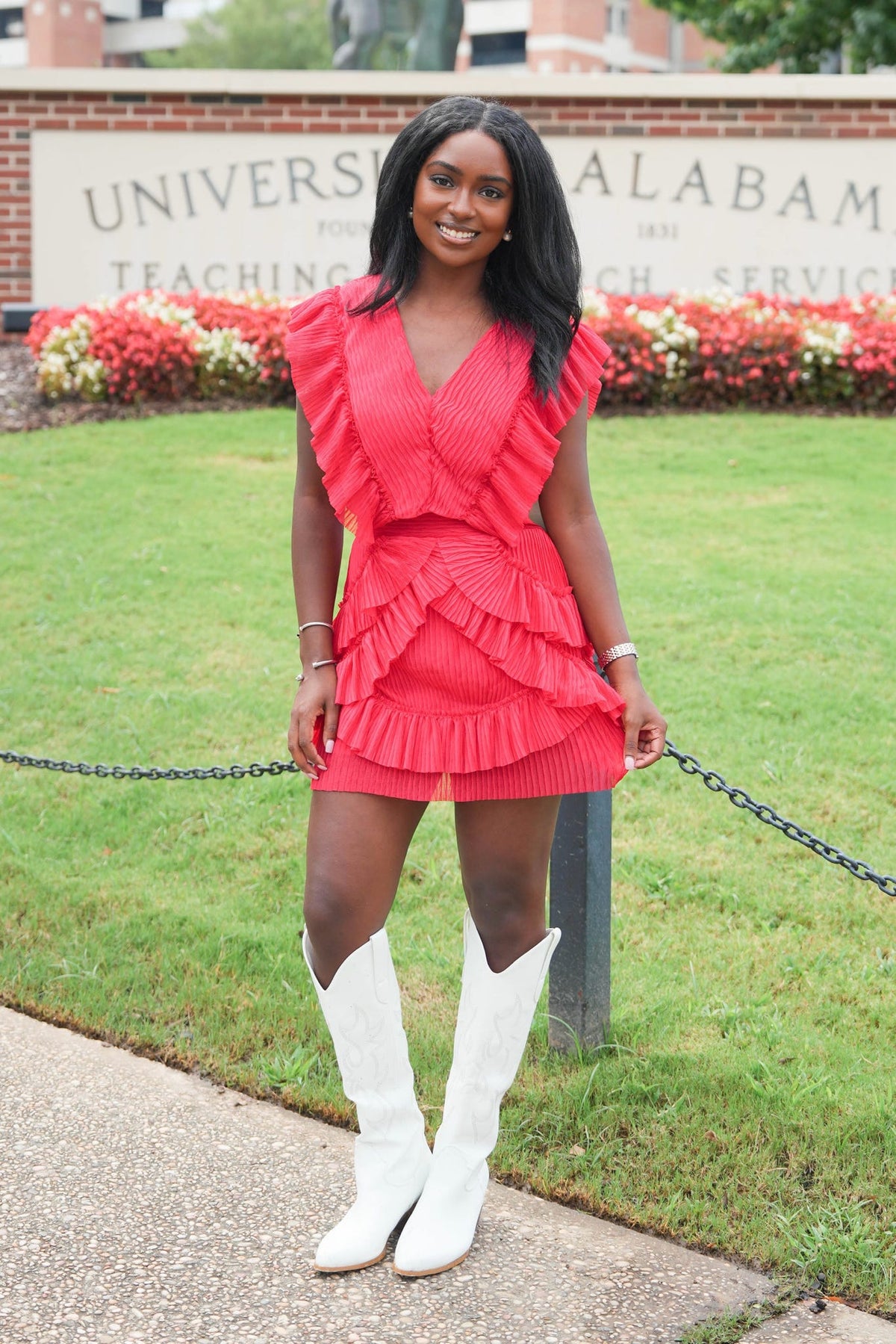 A woman stands outdoors on a paved walkway in front of a grassy lawn and a brick sign reading University of Alabama, wearing a bright coral-red ruffled wrap-style dress with white knee-high boots.