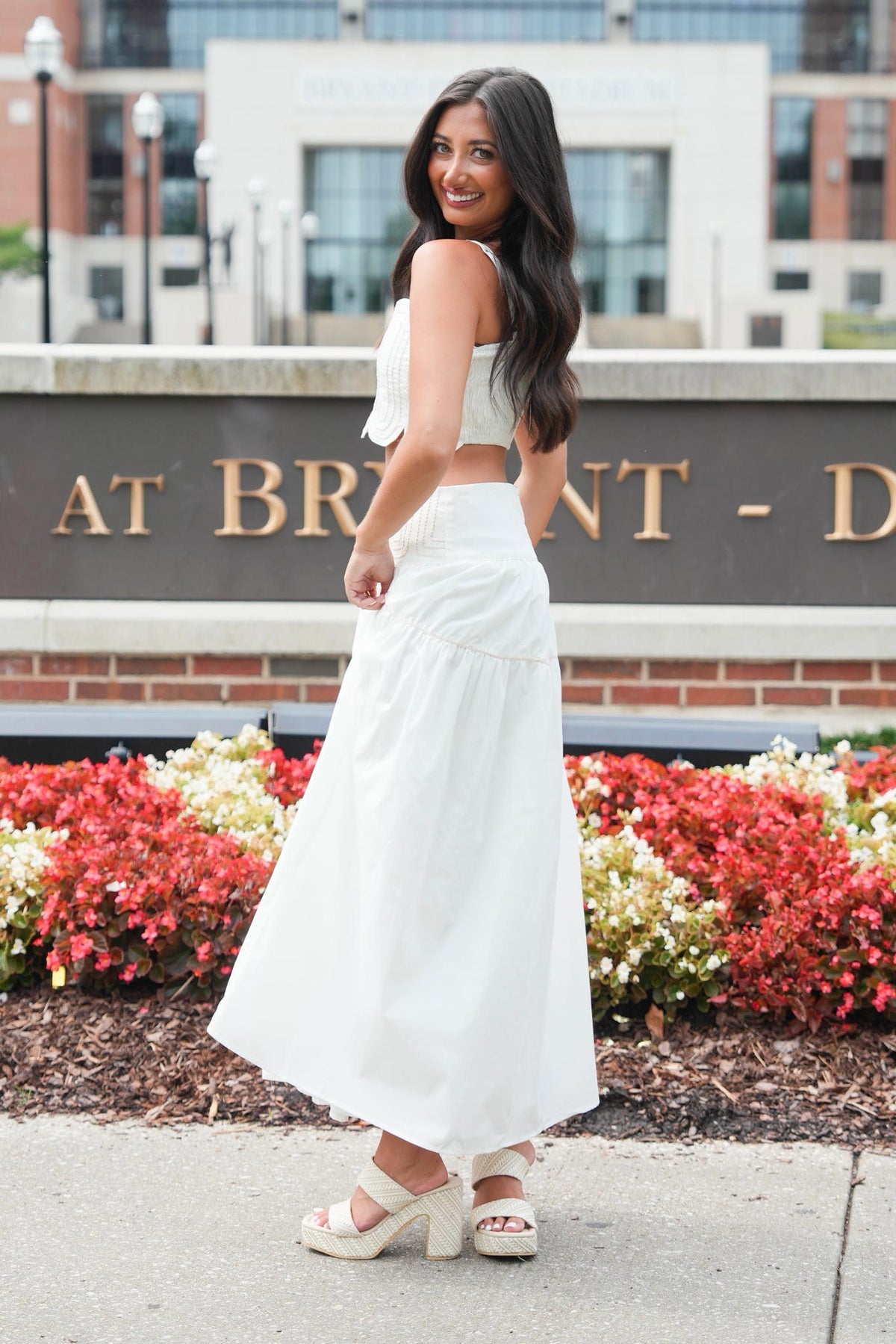 A woman wearing a white two-piece outfit consisting of a sleeveless crop top and a high-waisted maxi skirt, paired with white platform sandals, standing outdoors in front of a brick sign and flower beds.