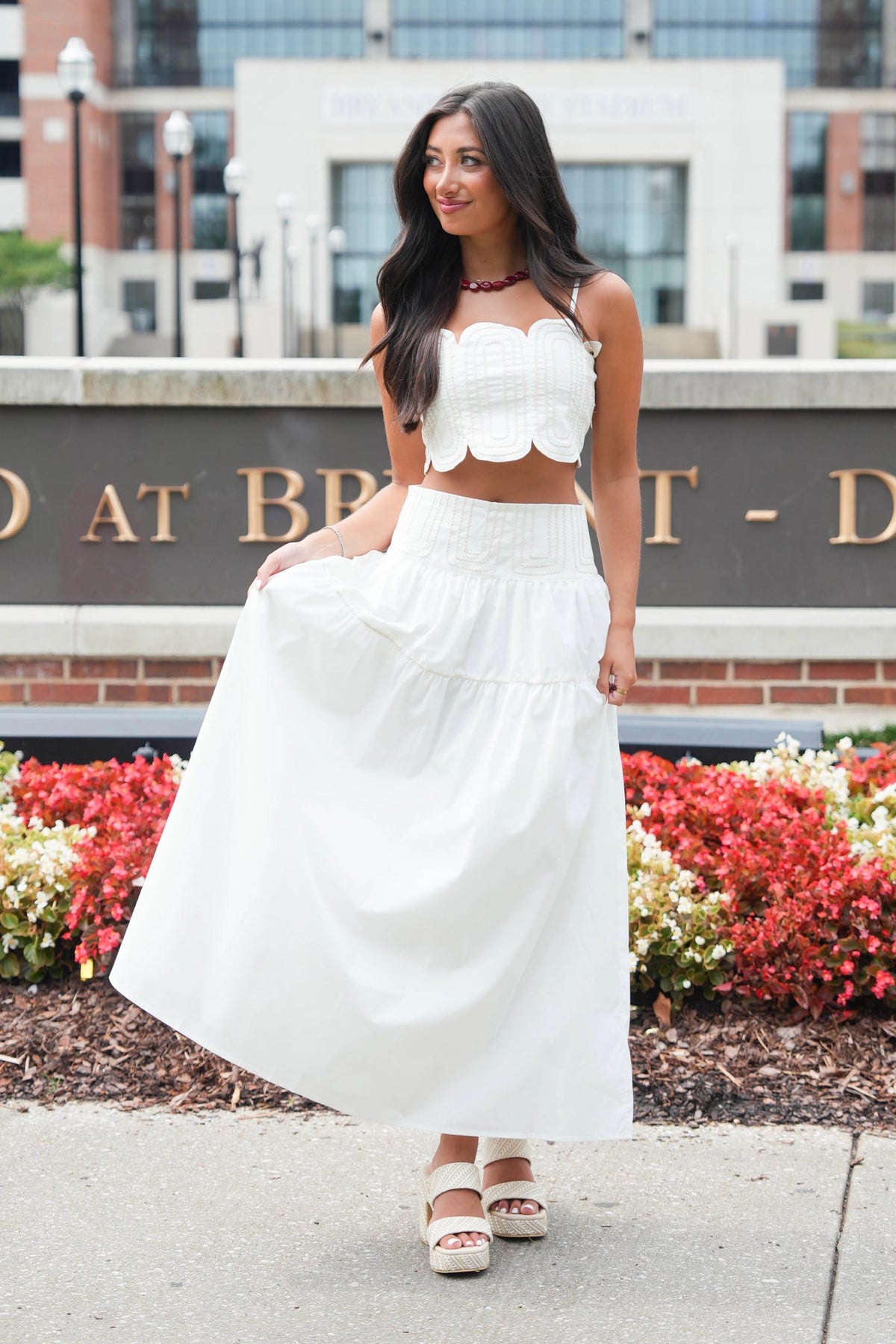 A woman modeling a white two-piece outfit consisting of a scalloped crop top and a matching tiered maxi skirt, standing outdoors in front of a brick building and flower beds.