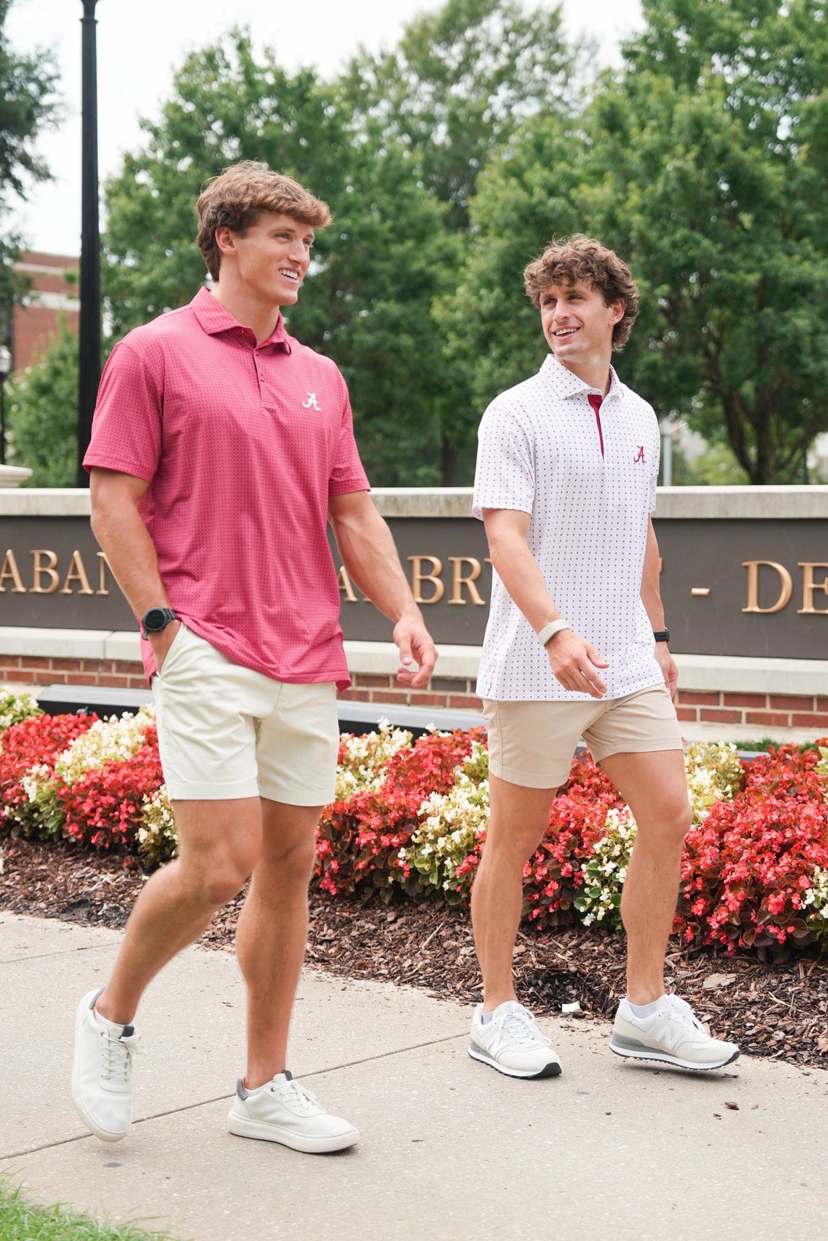 Two young men walking outdoors on a sidewalk in front of a brick sign and flower beds. The man on the left wears a pink polo shirt and beige shorts, while the man on the right wears a white patterned polo shirt and beige shorts.