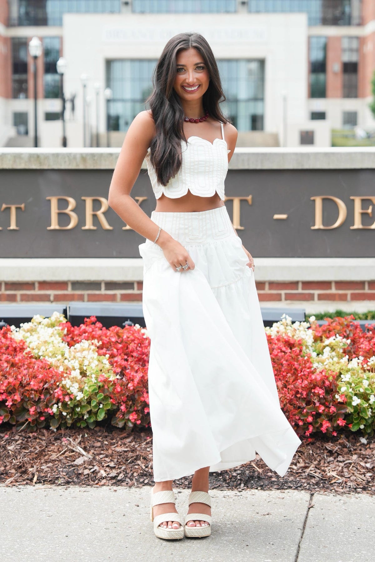 A woman modeling a white two-piece outfit consisting of a ruffled strapless crop top and a matching high-waisted maxi skirt, standing outdoors in front of a brick building and flower beds.