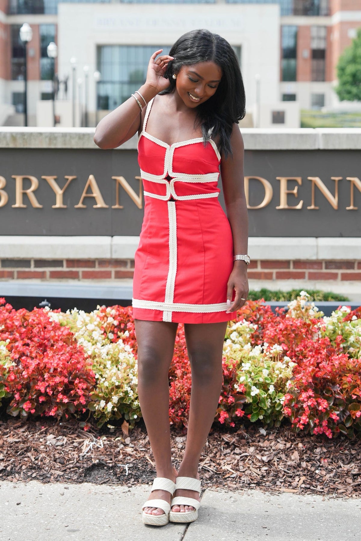 A woman modeling a red mini dress with white contrast trim and front cutout details, standing outdoors in front of a brick sign and flower beds.