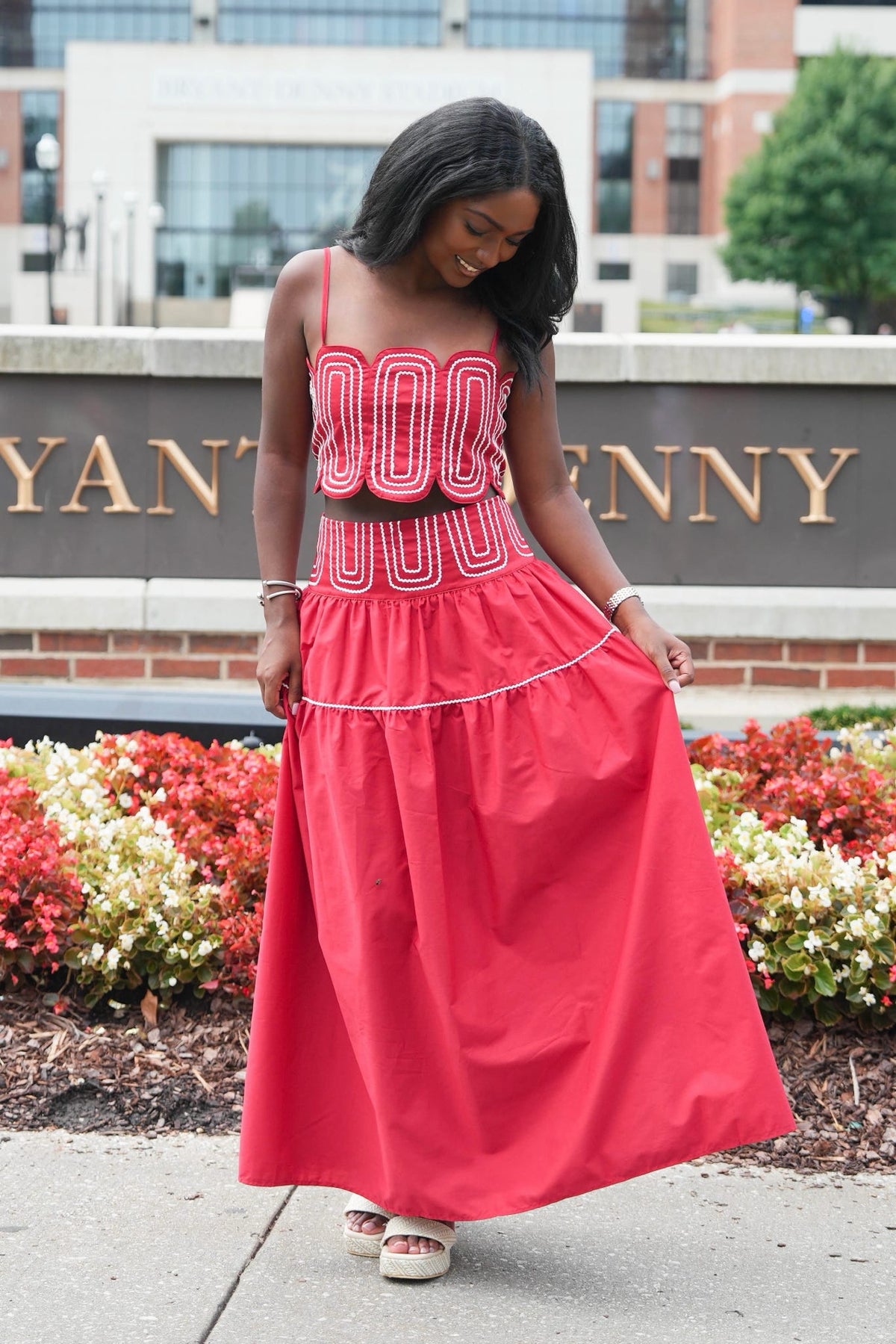 A woman wearing a bright coral pink two-piece dress with white geometric patterns on the bodice and a matching tiered skirt, standing outdoors in front of a brick sign with gold lettering.
