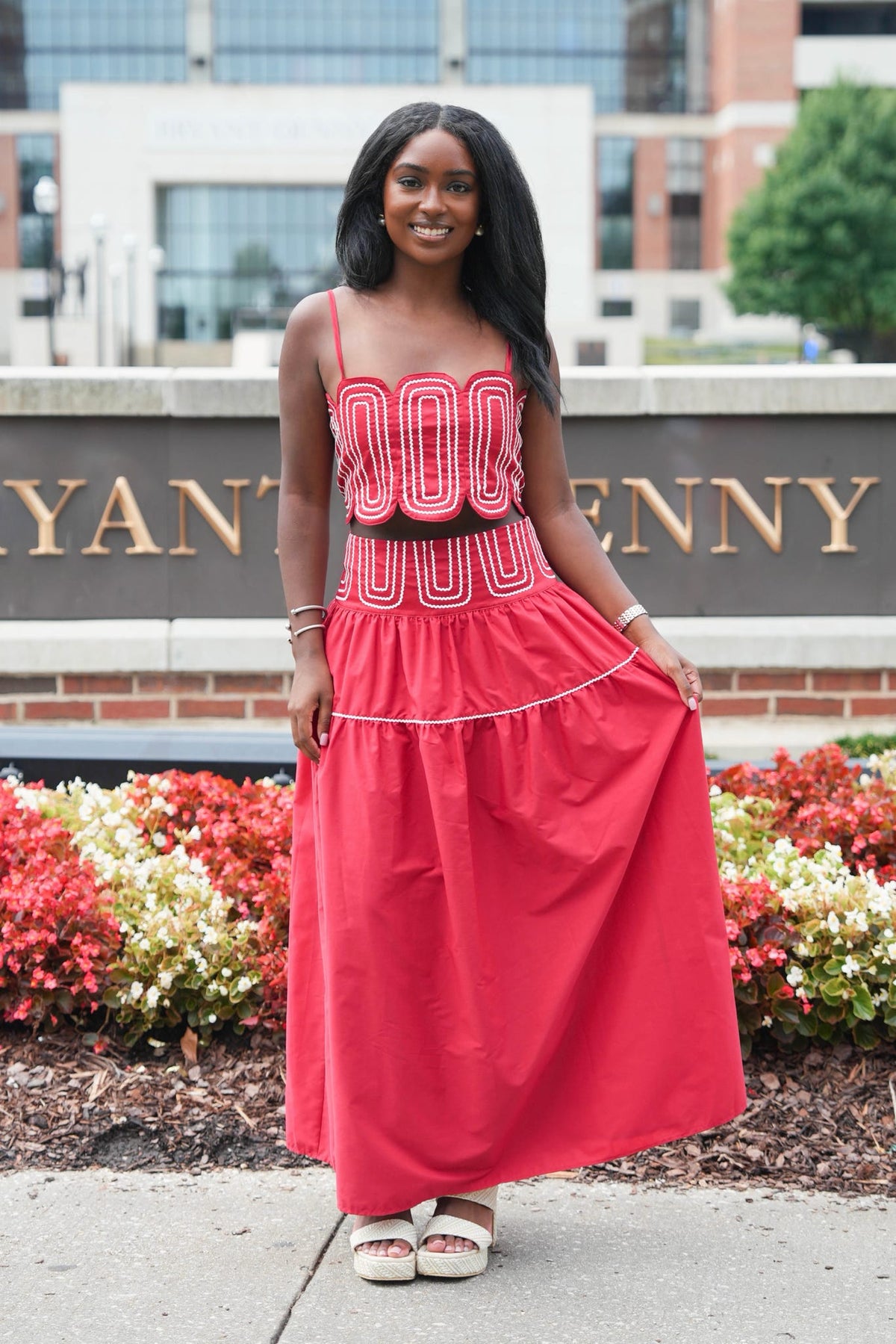 A woman modeling a bright red sleeveless maxi dress with white geometric patterns on the bodice and a matching red skirt, standing outdoors in front of a brick sign and flower beds.