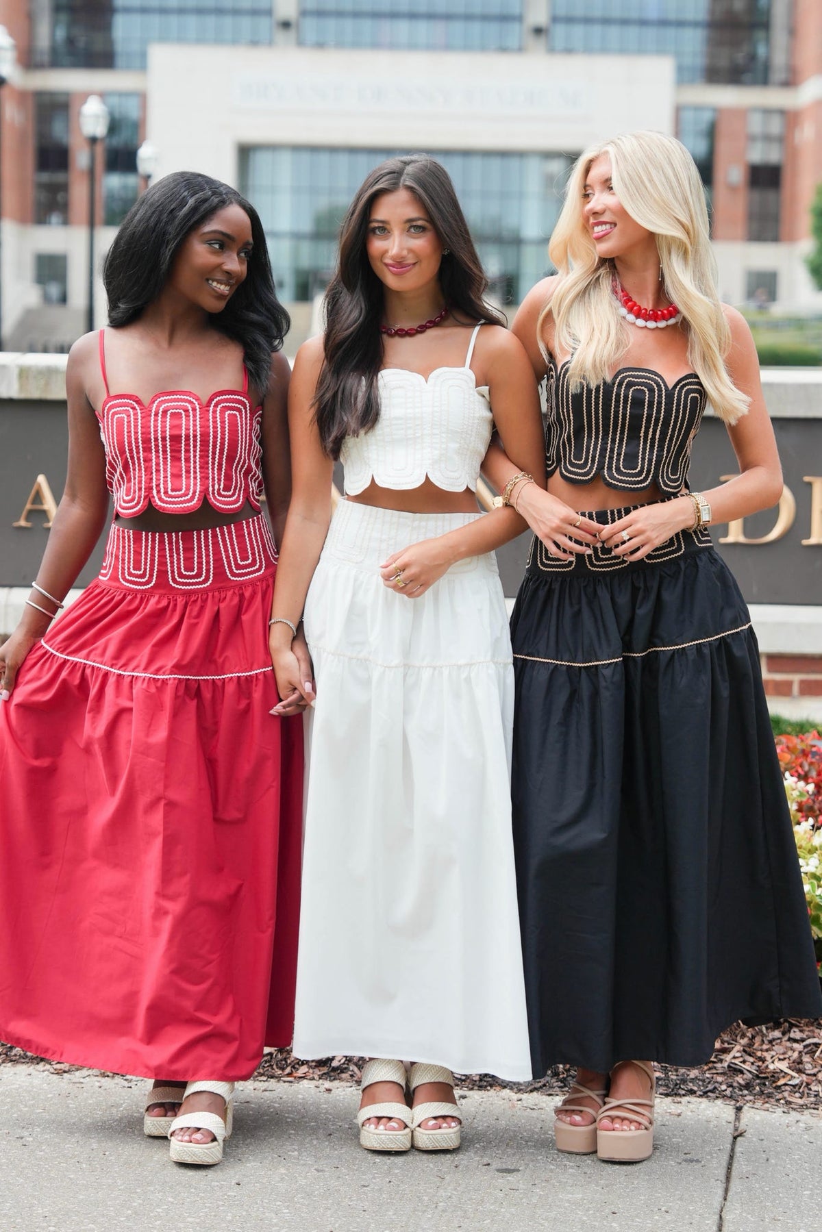 Three women standing outdoors wearing coordinated two-piece outfits with halter-style tops and long skirts. The outfits are in coral pink, white, and black, with decorative geometric patterns on the tops and contrasting trims on the skirts.