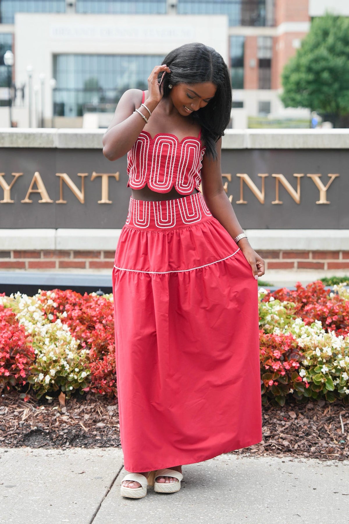 A woman wearing a bright pink two-piece matching set consisting of a strapless crop top with white geometric embroidery and a high-waisted, tiered maxi skirt, standing outdoors in front of a brick sign with gold lettering.
