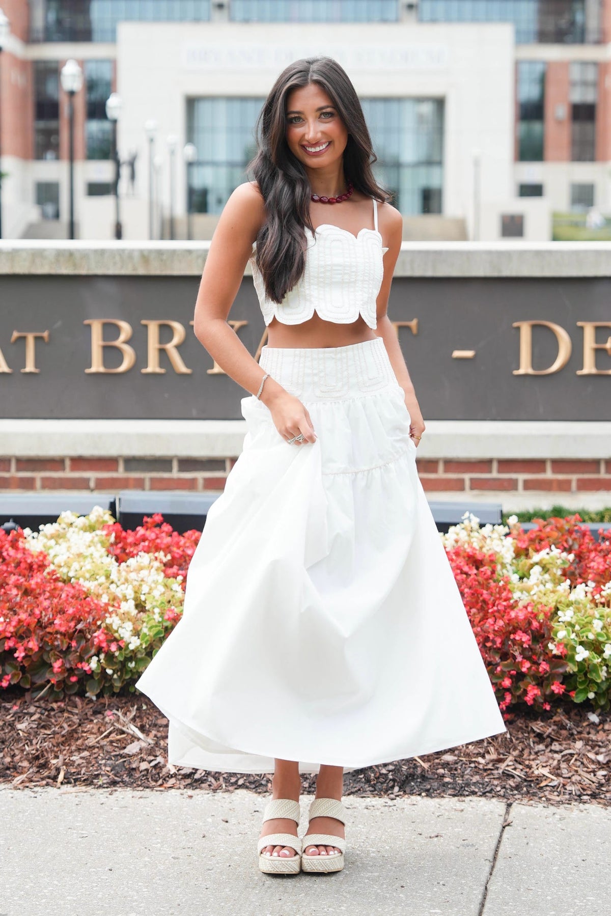 A woman modeling a white two-piece outfit consisting of a ruffled spaghetti-strap crop top and a matching high-waisted tiered maxi skirt, paired with white platform sandals, set against a brick and stone background with flowers.