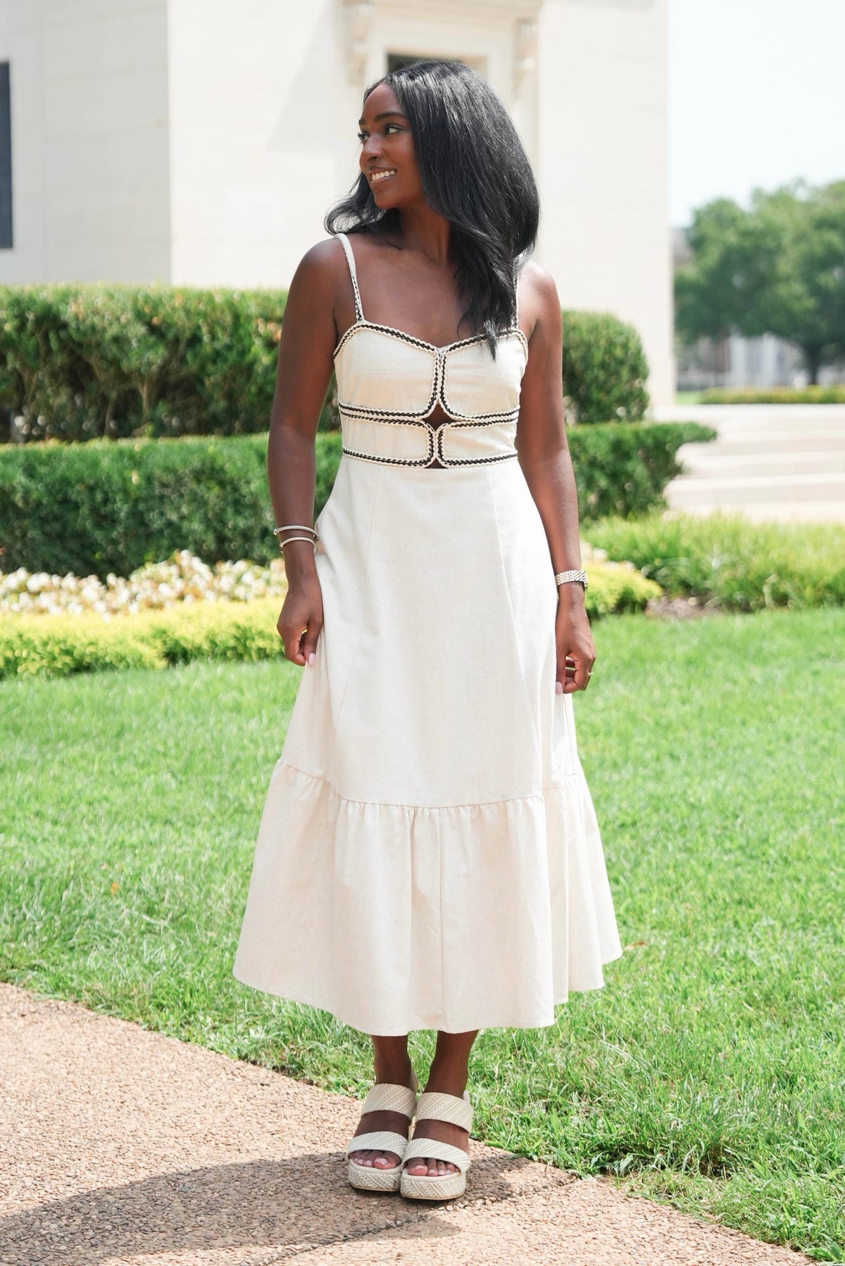 A woman wearing a cream-colored sleeveless midi dress with a tiered skirt and decorative gold trim, standing outdoors on a paved path with a green lawn and building in the background.