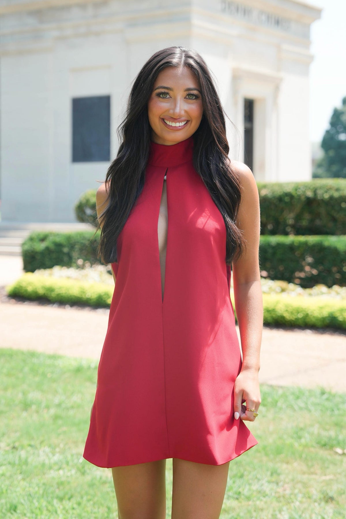 A woman with long dark hair stands outdoors in front of a light-colored building and green hedges, wearing a sleeveless red dress with a high mock neck and a front keyhole cutout.
