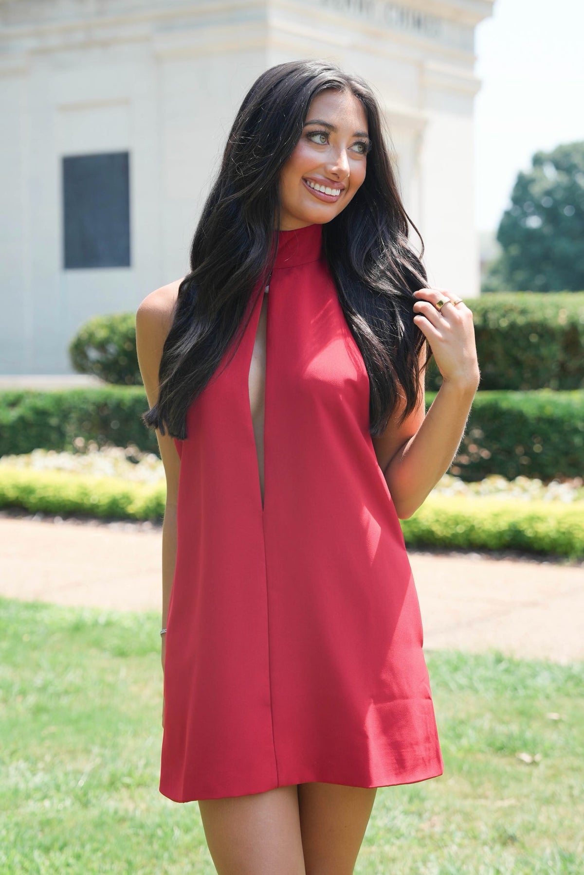 A woman with long dark hair wearing a sleeveless red dress with a high mock neck and front slit, standing outdoors in a grassy park area with a building in the background.