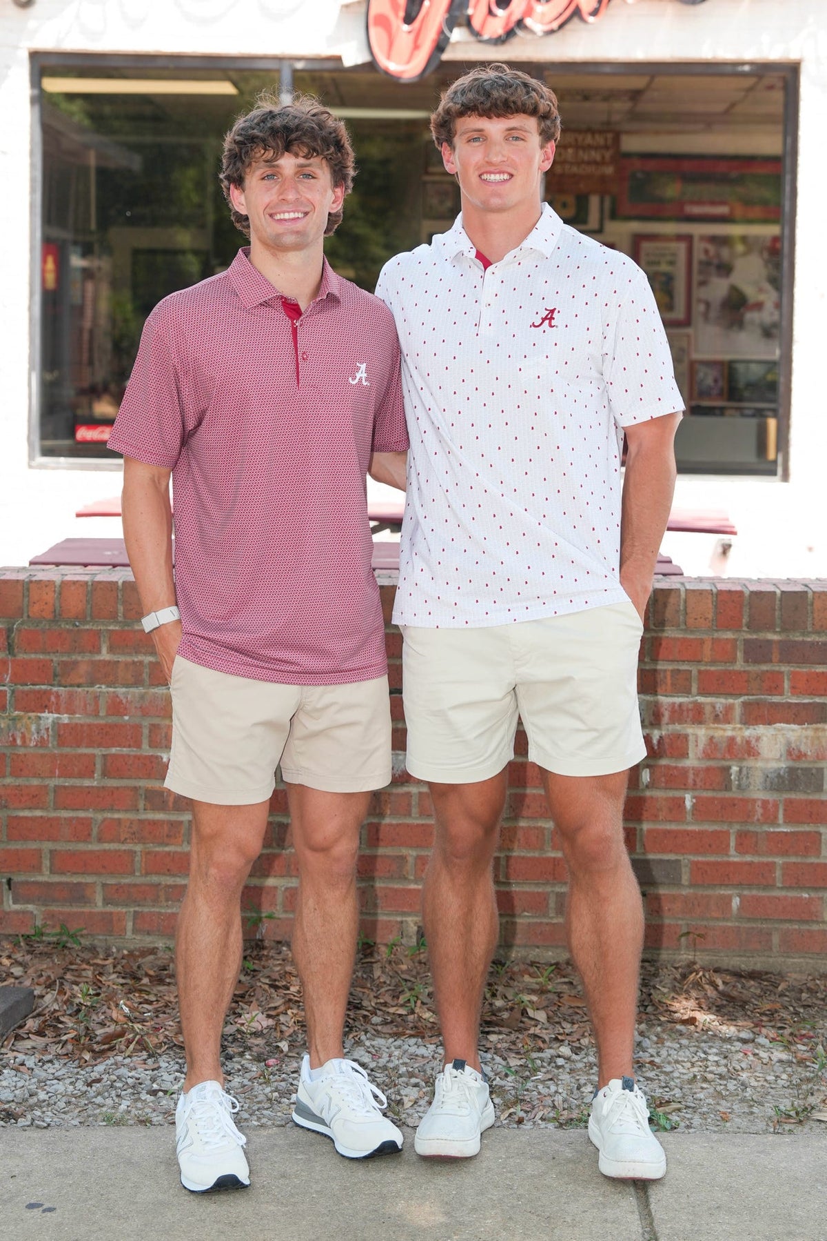 Two young men standing outdoors in front of a brick building, wearing polo shirts and shorts. The man on the left wears a dusty rose polo shirt with a red collar and a red 'A' logo on the chest, paired with khaki shorts and white sneakers.
