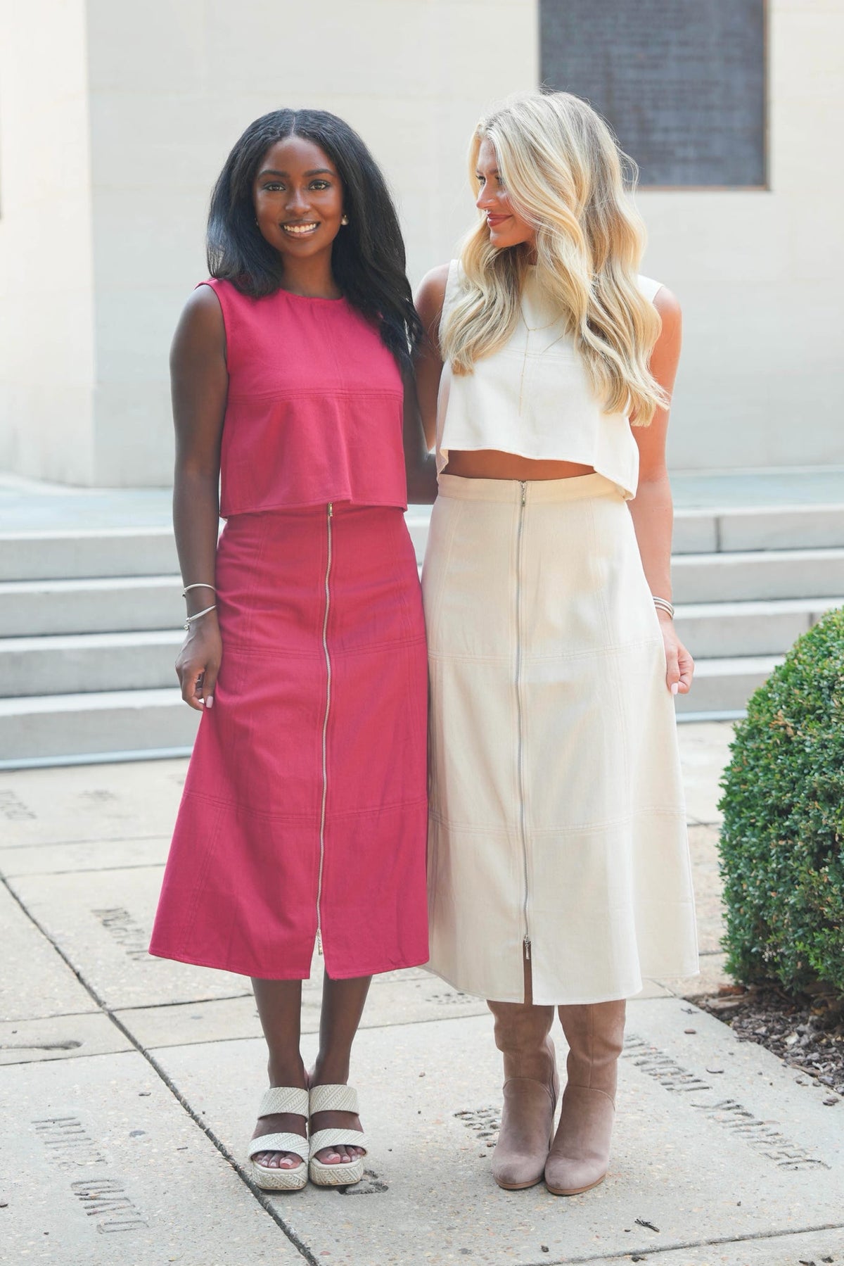 Two women standing outdoors in front of a building entrance. The woman on the left wears a sleeveless bright pink two-piece outfit with a ruffled top and midi skirt.