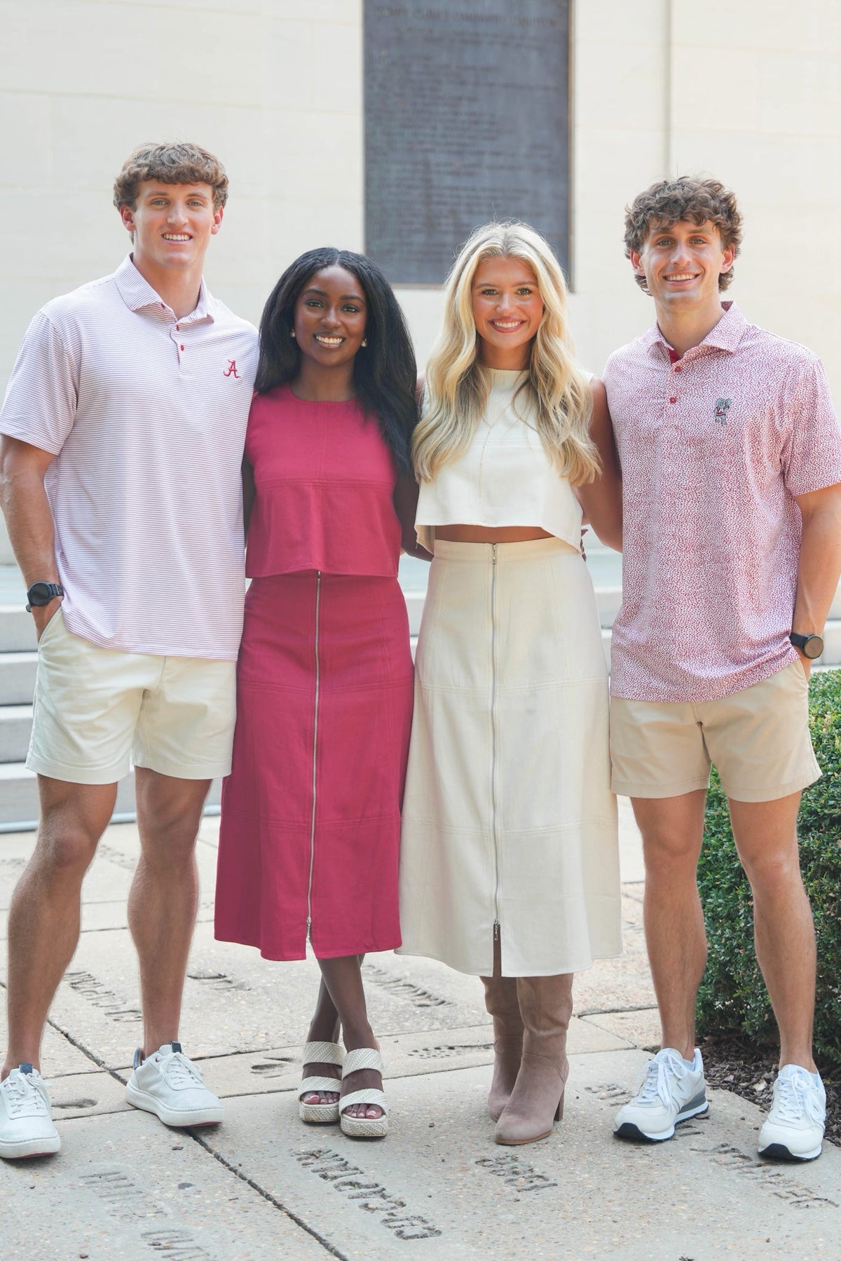 Four young adults standing together outdoors in front of a light-colored building. Two wear light pink polo shirts with beige shorts, and two wear a matching pink and cream set with a white crop top and cream skirt.