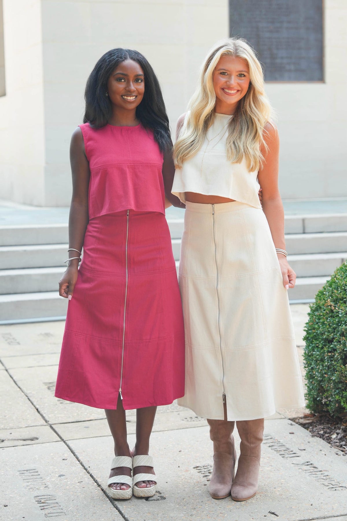Two women standing outdoors in front of a building entrance. The woman on the left wears a bright pink sleeveless midi dress with a front zipper, and the woman on the right wears a cream sleeveless crop top with a matching high-waisted midi skirt and beige knee-high boots.