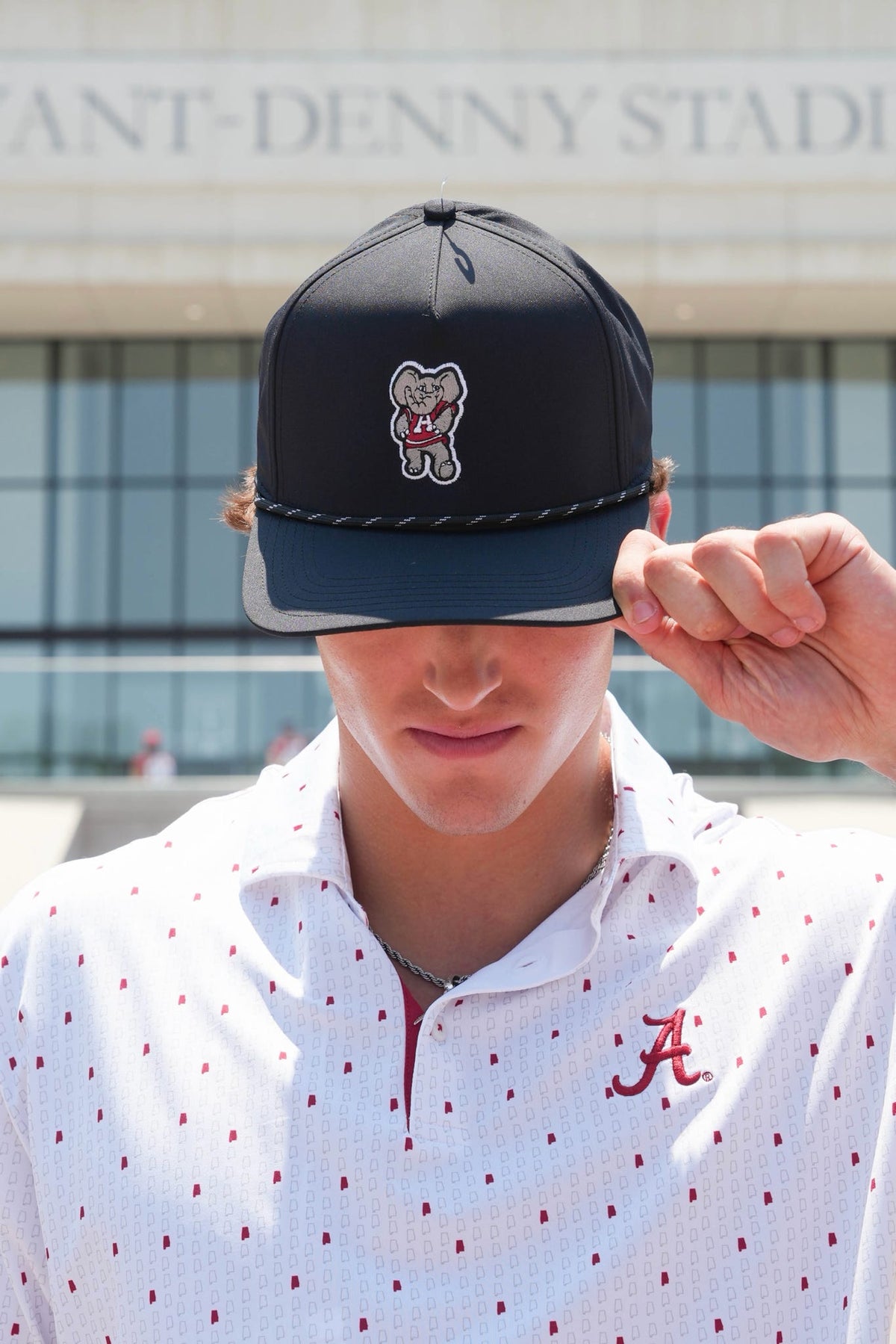 A man wearing a dark navy blue baseball cap with a white embroidered bear logo and a white polo shirt with red polka dots and a red 'A' logo, standing outdoors in front of a stadium building.