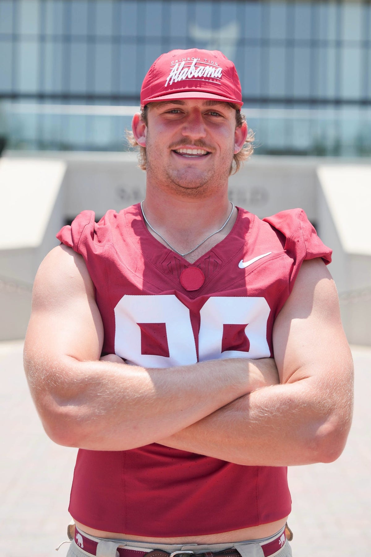 A smiling man wearing a crimson red Alabama football jersey with the number 00 and a matching red cap stands with his arms crossed. The Nike swoosh is visible on the chest, and the background shows a modern glass building.