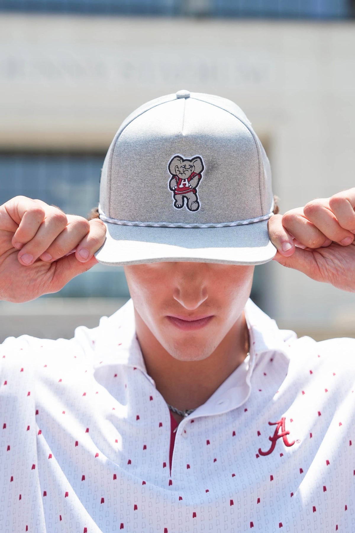 A person wearing a light gray snapback cap with a cartoon bear patch on the front, holding the brim down over their eyes. They are wearing a white polo shirt with red dots and a red 'A' logo on the chest.
