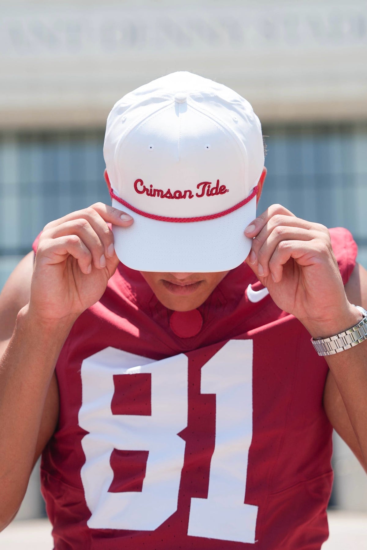 A person wearing a white baseball cap with 'Crimson Tide' in red script and a matching red athletic jersey with the number 81, holding the brim of the cap in front of their face.