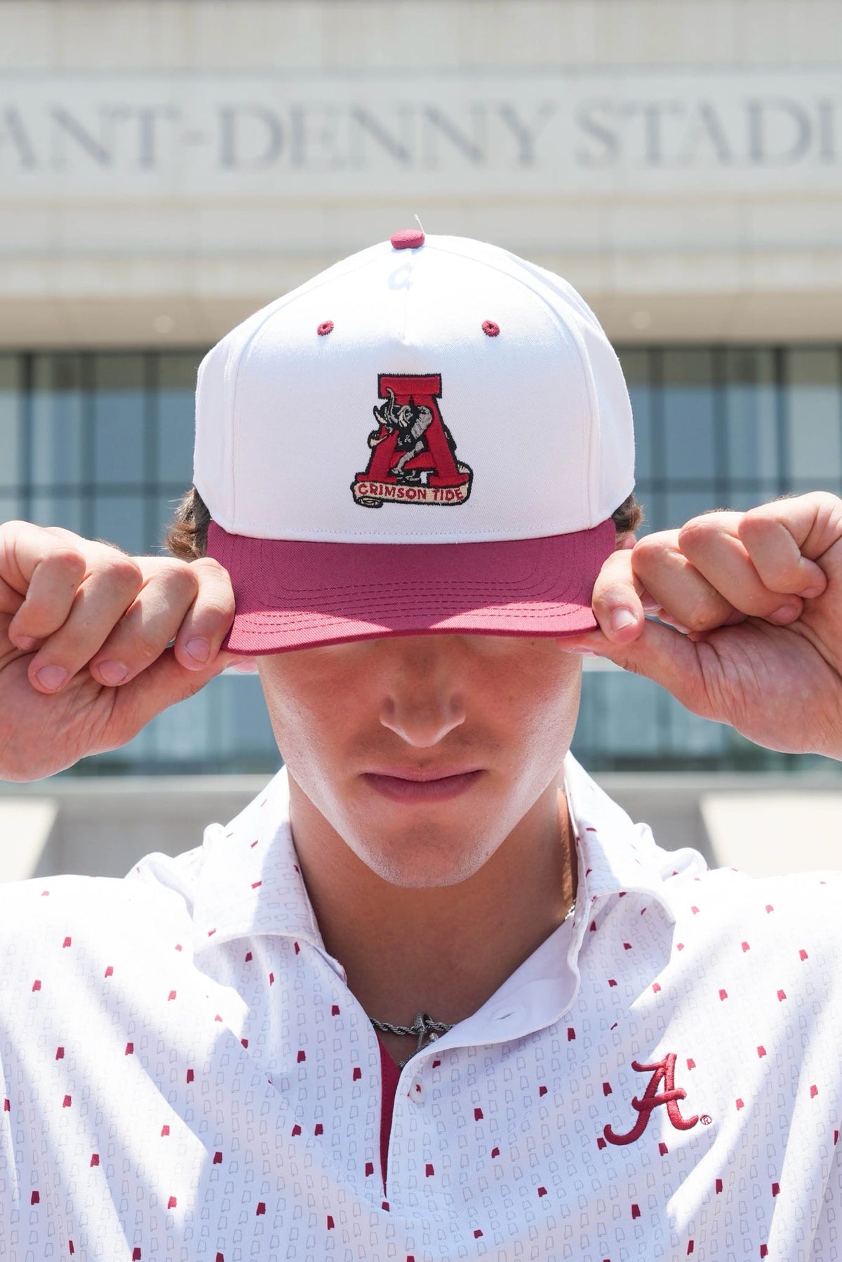 A person outdoors wearing a white and maroon Alabama Crimson Tide snapback cap, holding the brim down over their eyes. The cap features a large crimson 'A' logo with a bear mascot on the front.