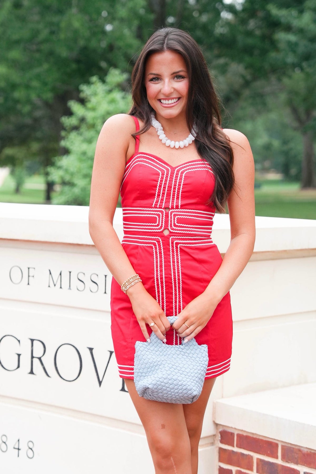 A woman wearing a red and white striped mini dress, holding a light blue textured handbag, standing outdoors in front of a stone wall with green trees in the background.