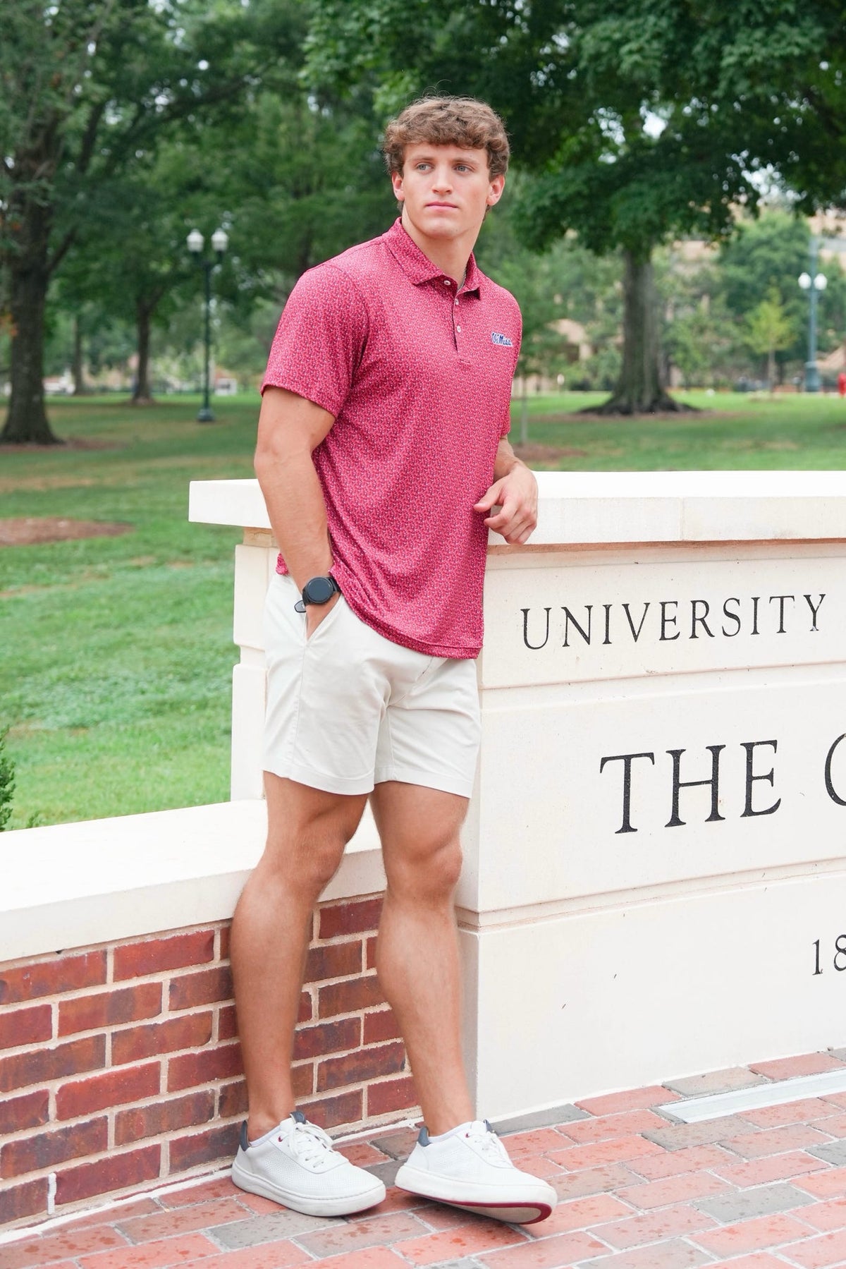 A young man stands outdoors leaning against a white stone wall with brick steps below, wearing a short-sleeve textured polo shirt in heathered red, white shorts, and white sneakers. Trees and grass are visible in the background, and part of a sign reading 'UNIVERSITY THE C.