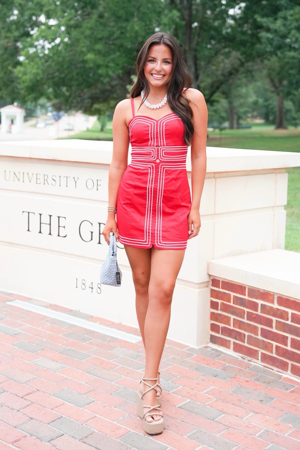 A woman stands outdoors wearing a bright red sleeveless mini dress with white geometric stripe details, holding a small gray handbag. The background shows a stone wall with University of Georgia lettering and trees.