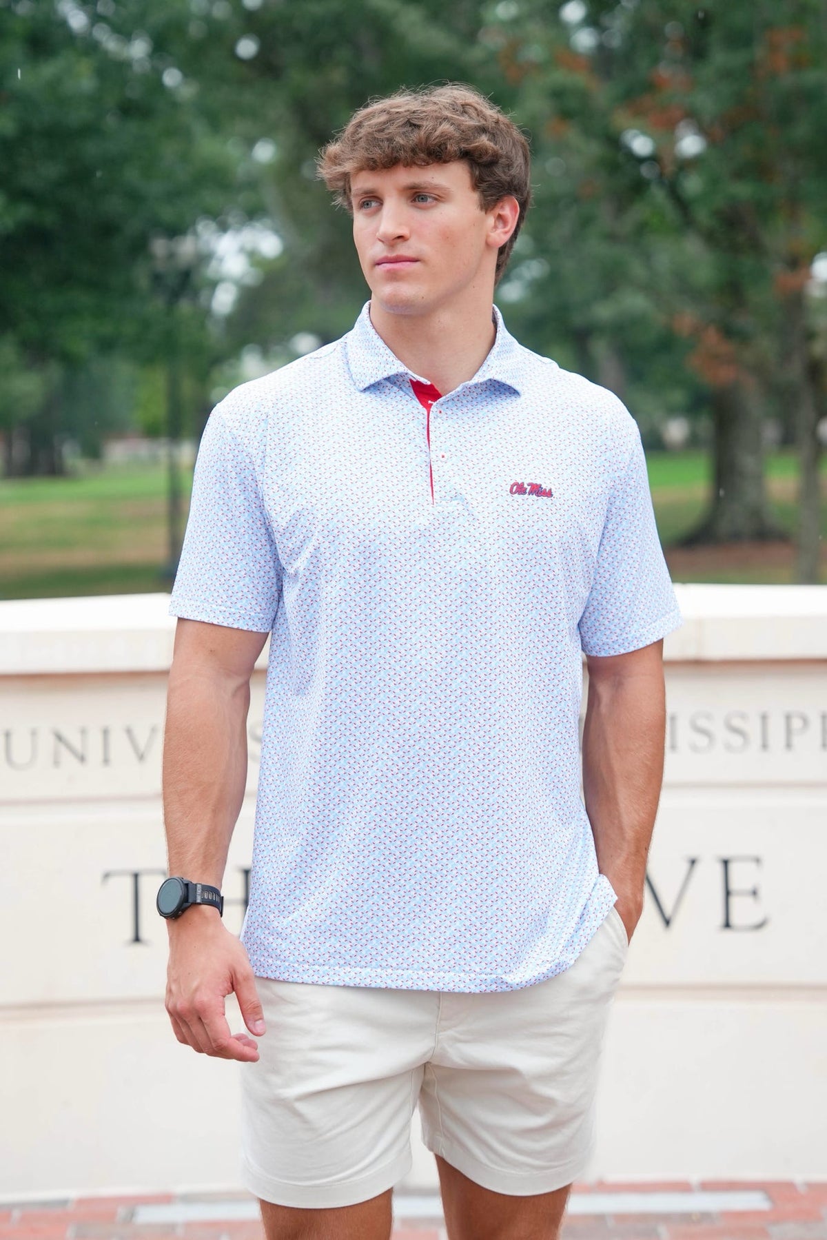 A young man wearing a light blue and white patterned short-sleeve polo shirt with a red zipper and a small red logo on the chest, paired with beige shorts, standing outdoors in front of a stone wall with trees in the background.