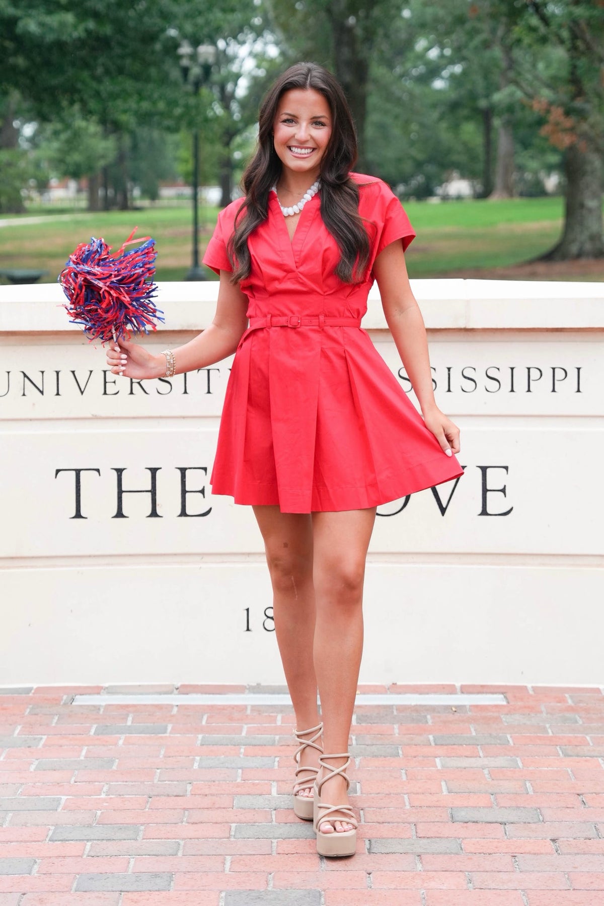 A woman stands outdoors in front of a University of Mississippi sign, wearing a bright red short-sleeve mini dress with a flared skirt and holding a red, white, and blue pom-pom.