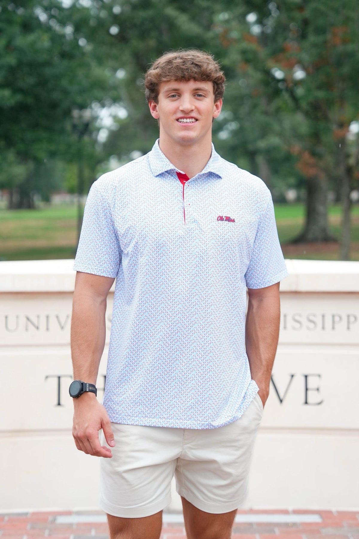 A young man smiling outdoors wearing a light blue and white patterned polo shirt with a red zipper and a small red logo on the chest, paired with beige shorts.
