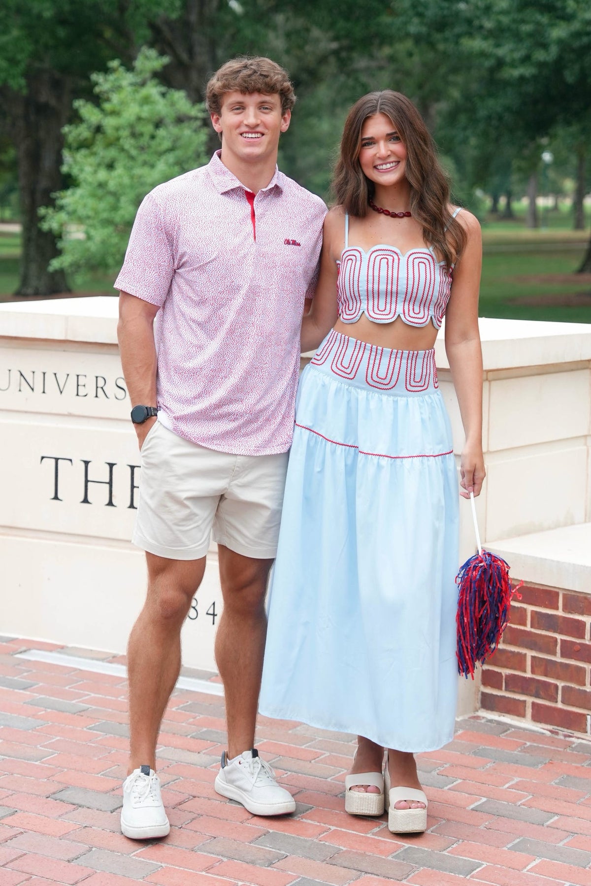 A young man and woman standing together outdoors on a brick walkway in front of a stone wall with trees in the background. The man wears a light pink polo shirt, white shorts, and white sneakers, while the woman wears a light blue sleeveless crop top and matching long skirt with red trim, along with white platform sandals and holds a red and blue pom-pom.