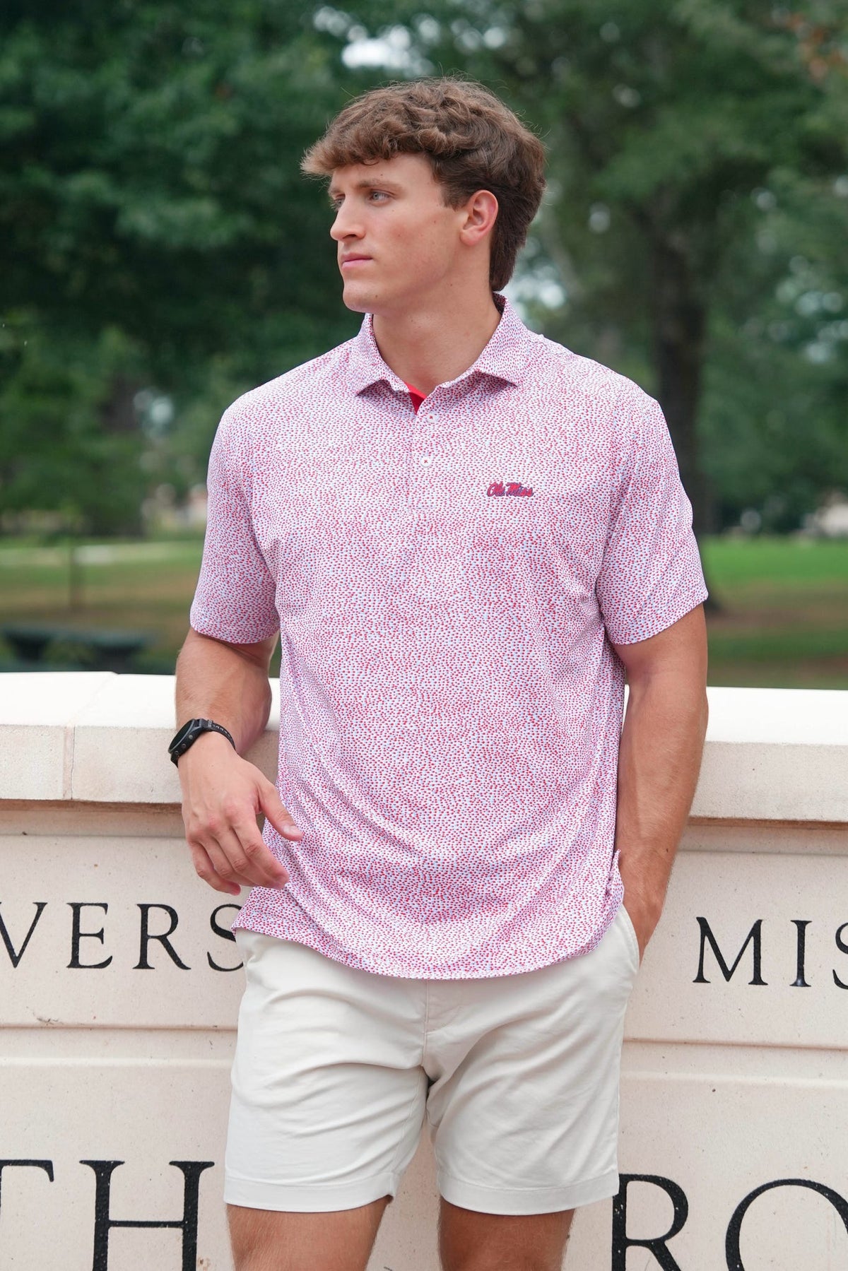 A man wearing a light pink and white patterned short-sleeve polo shirt with white shorts, standing outdoors in front of a stone wall with green trees in the background.